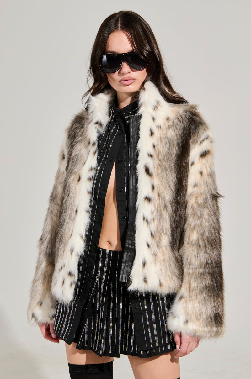 A person with long dark hair wears oversized black sunglasses and the chic ELSIE FAUX FUR BOMBER, featuring a luxurious animal print design, over a partially unbuttoned black shirt and pinstriped shorts. The plain, neutral background emphasizes their stylish ensemble.