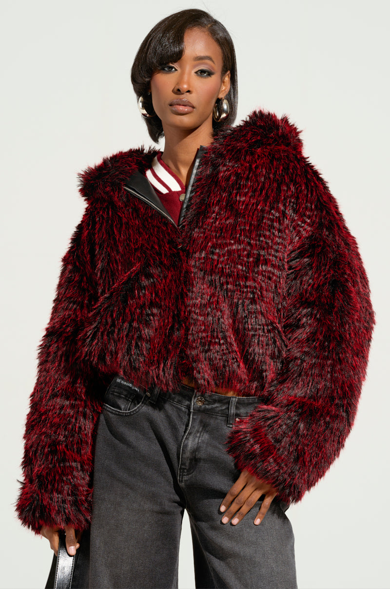 A person is wearing the NIKKI FAUX FUR BOMBER in red and black, featuring a metallic silver zip, with a maroon top underneath. The ensemble is completed with dark jeans. The individual has short hair and is posing against a plain background while holding a bag in one hand.
