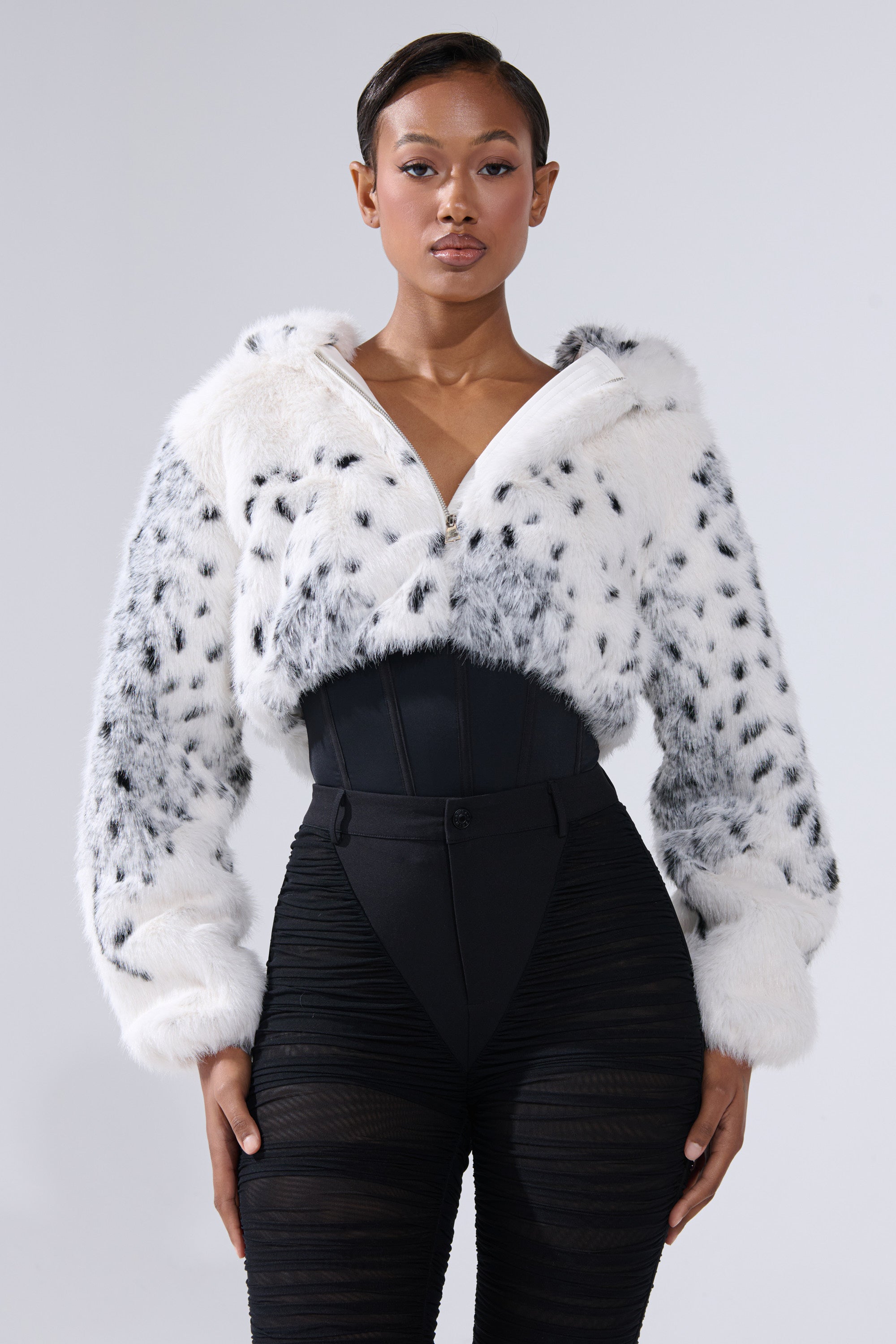 A woman faces forward in the SNOW LEOPARD HOODED FUR BOMBER with black spots, paired with a black high-waisted corset belt and sheer black pants. The background is plain light gray.