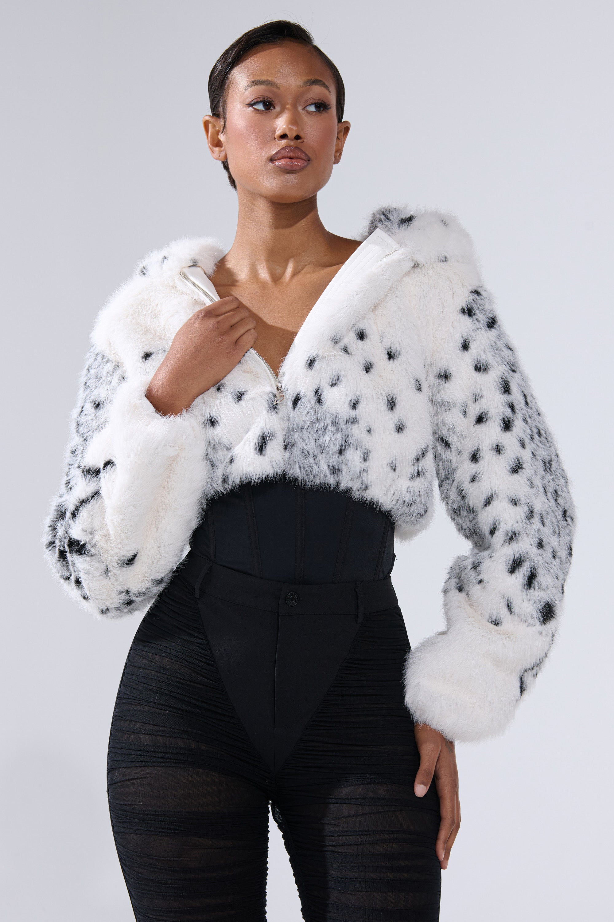 A woman poses confidently against a plain light background, wearing the SNOW LEOPARD HOODED FUR BOMBER with black spots and a hood, styled with high-waisted black pants and a sleek black corset.