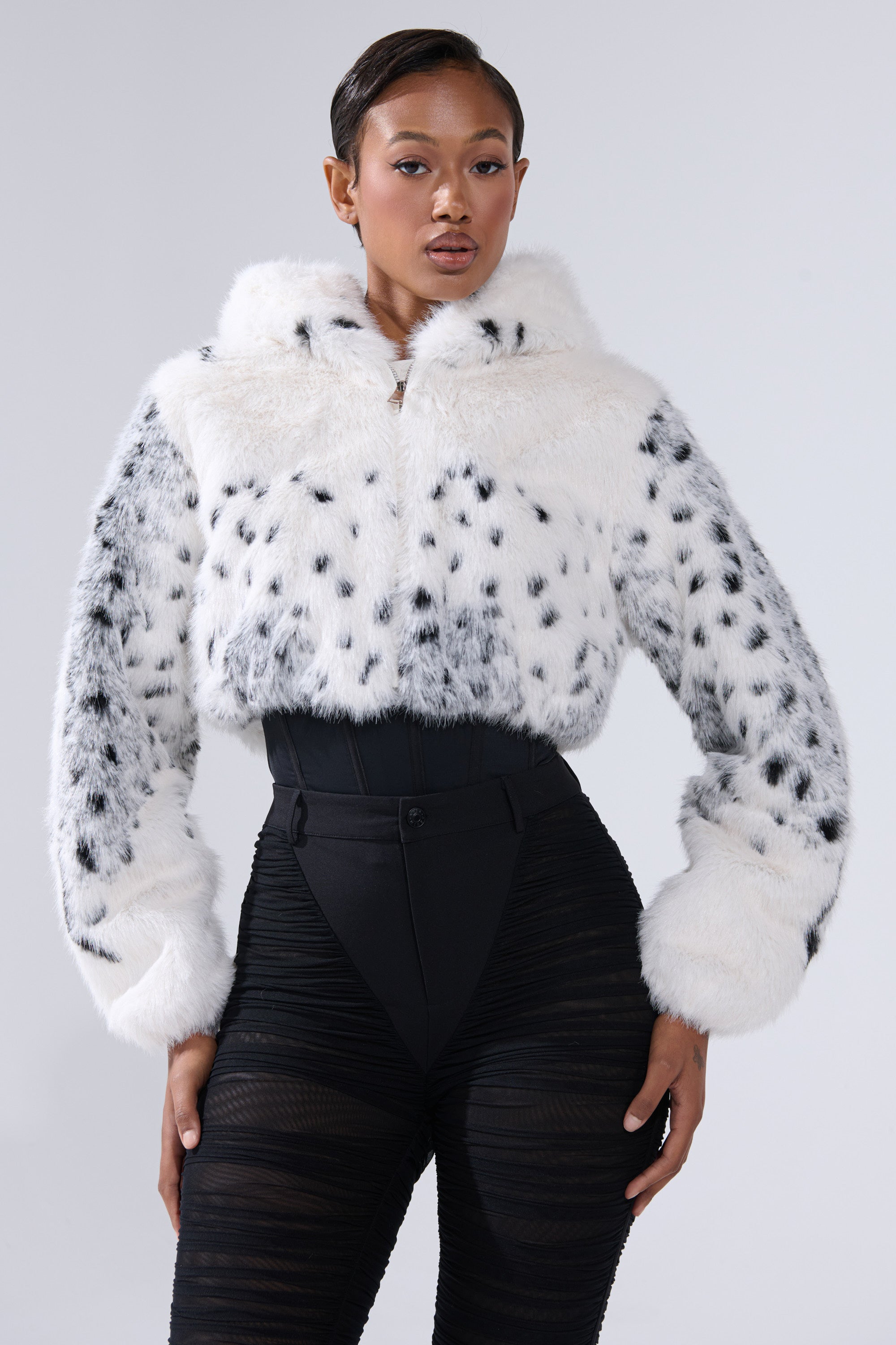 A woman models the SNOW LEOPARD HOODED FUR BOMBER with black spots and hood, styled with high-waisted black sheer leggings, posing confidently against a plain light background.