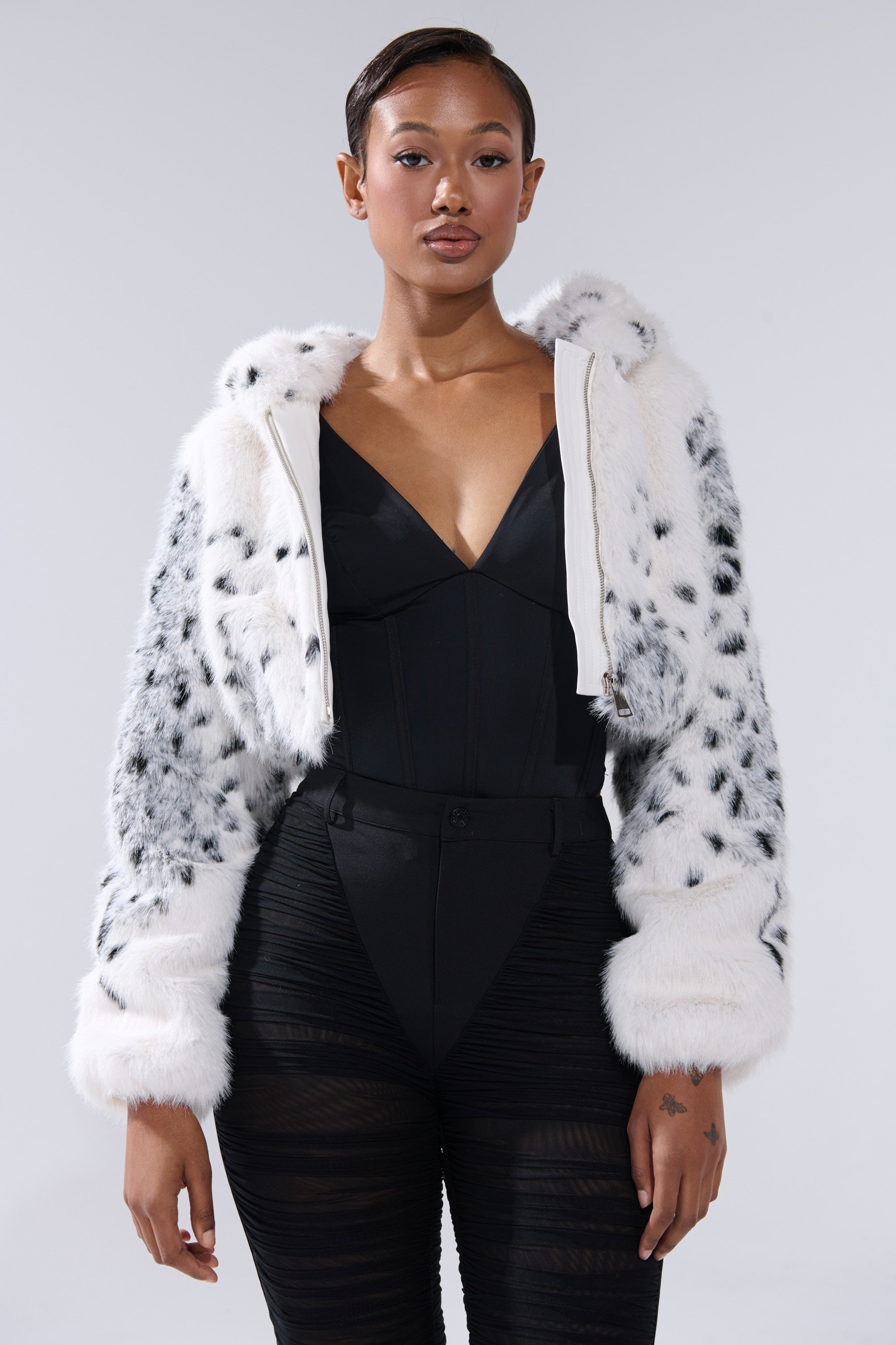 A woman with short hair stands confidently against a plain light background, wearing a black bodysuit and the SNOW LEOPARD HOODED FUR BOMBER.