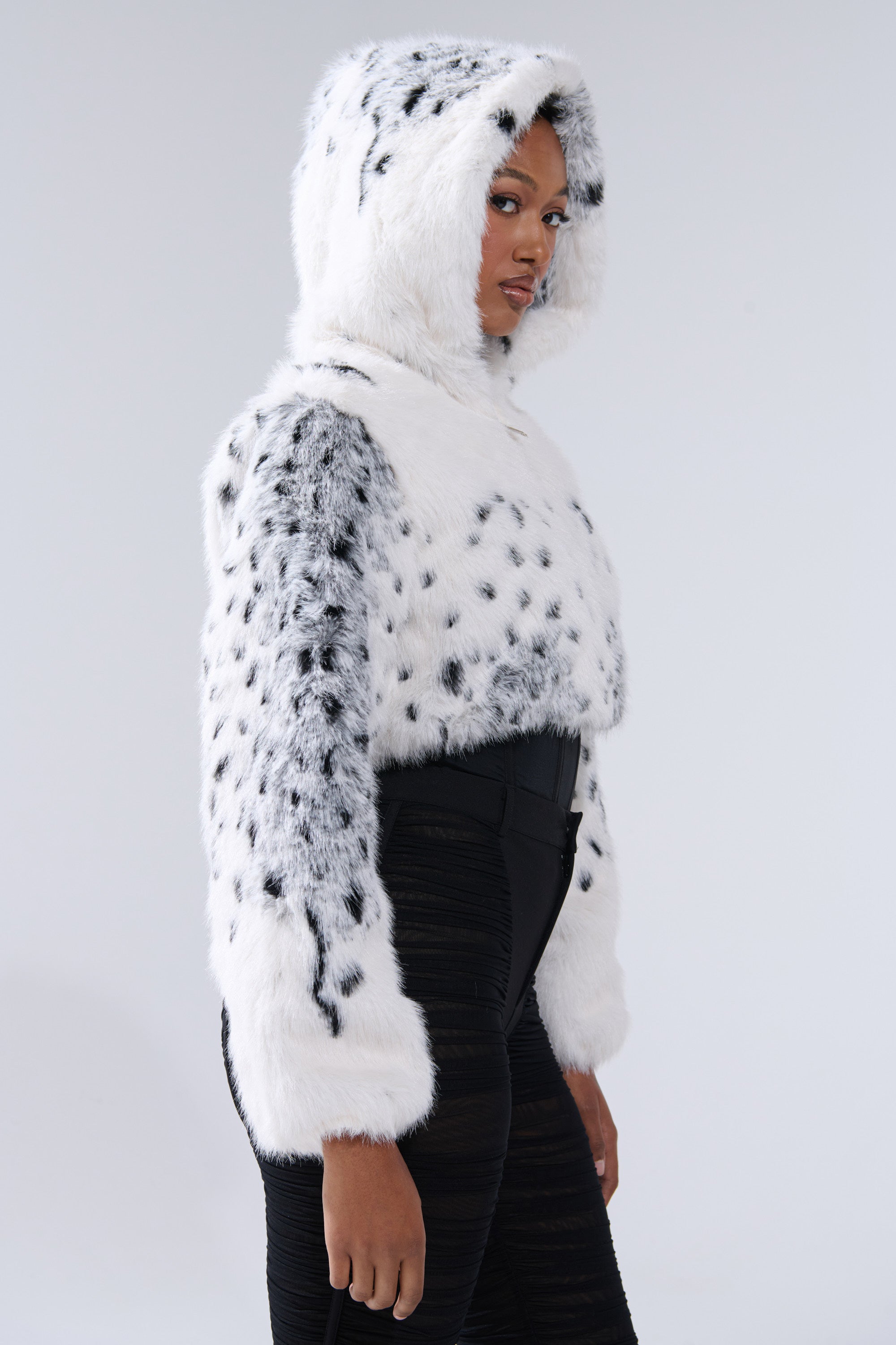 Wearing the SNOW LEOPARD HOODED FUR BOMBER with black spots and black high-waisted pants, a person stands against a plain light background, gazing to the side.