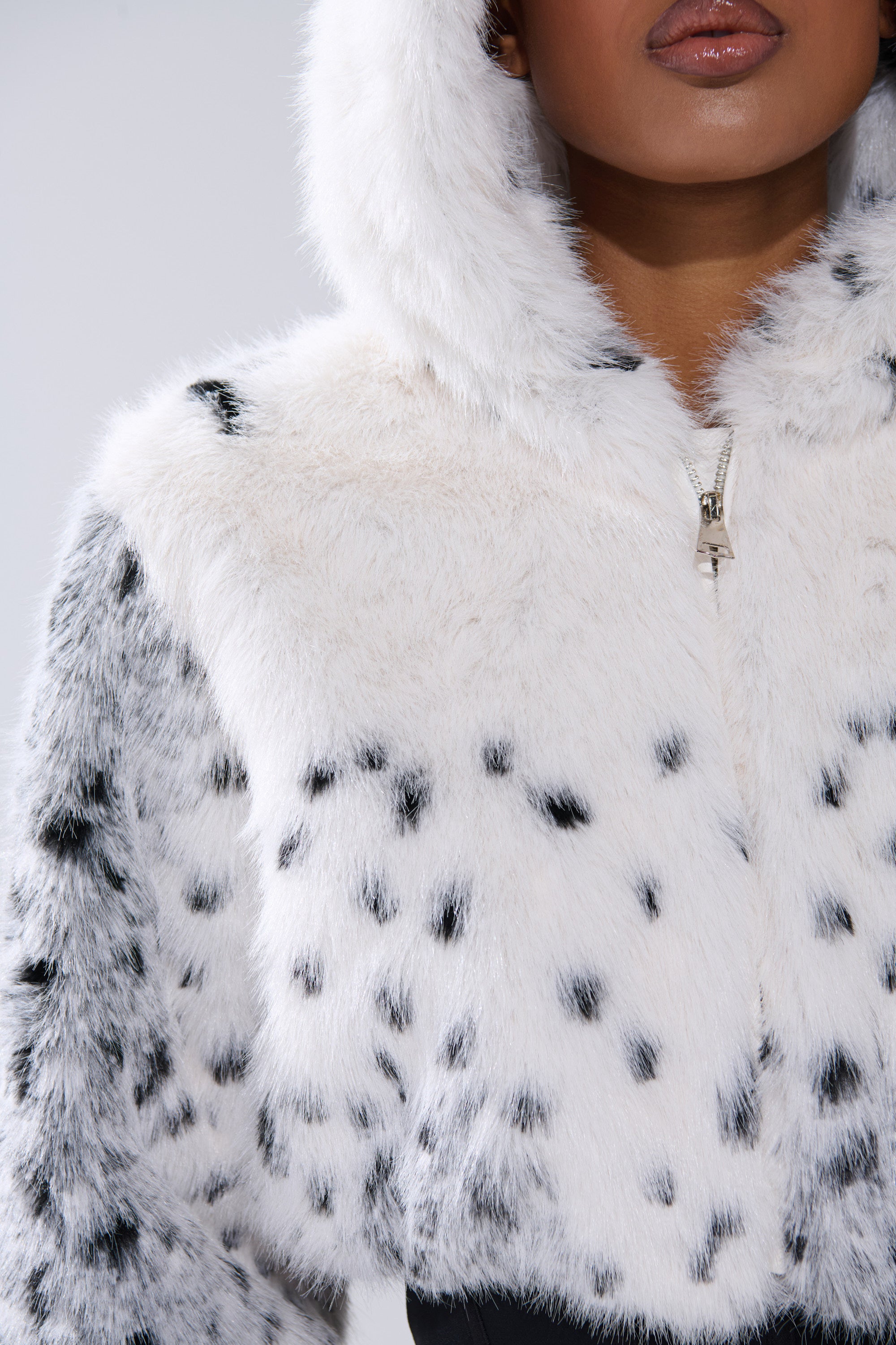 Wearing a SNOW LEOPARD HOODED FUR BOMBER with white faux fur and black spots, hood partially zipped, the focus is on the upper body while eyes remain out of frame.