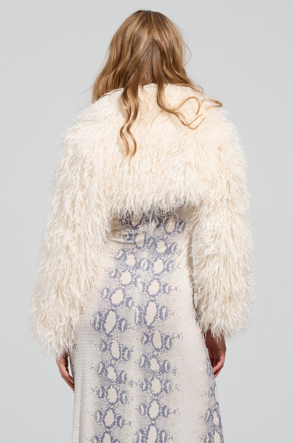 The individual with long hair is donned in sophisticated outerwear—a chic LILIANA ULTRA CROP FAUX FUR jacket—standing with their back to the camera. Perfectly complemented by sleek stiletto booties and a long, patterned snakeskin dress featuring light and dark gray tones, their ensemble radiates contemporary elegance.