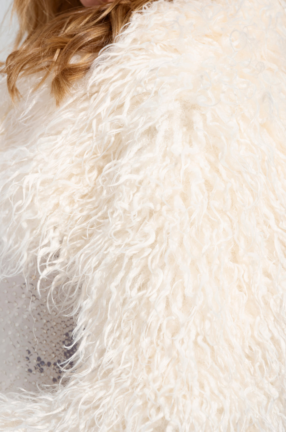 Close-up of a person in chic outerwear, showcasing the LILIANA ULTRA CROP FAUX FUR coat in white. Blonde hair peeks out at the top as the coat's curly and soft texture dominates the image's upper part.