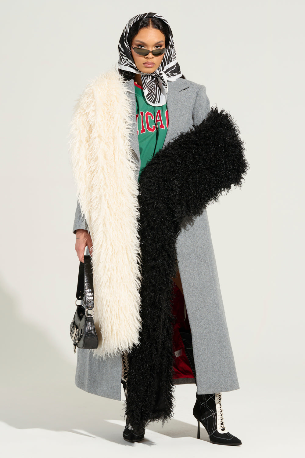 A stylish individual flaunts a long gray coat layered over a red and green color block pattern shirt, complemented by sunglasses and a leopard scarf. They carry the EPIC DREAMS FAUX FUR SCARF with its polyester lining and clutch a small handbag, while their zebra-print boots contrast strikingly against the plain background.
