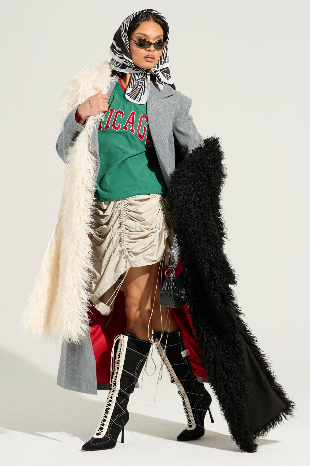 A stylish person poses confidently in a layered outfit, featuring a gray coat with black and white fur trims, accentuated by the elegant EPIC DREAMS FAUX FUR SCARF. They wear a green "Chicago" shirt, a ruched beige skirt, lace-up boots, sunglasses, and a patterned headscarf.