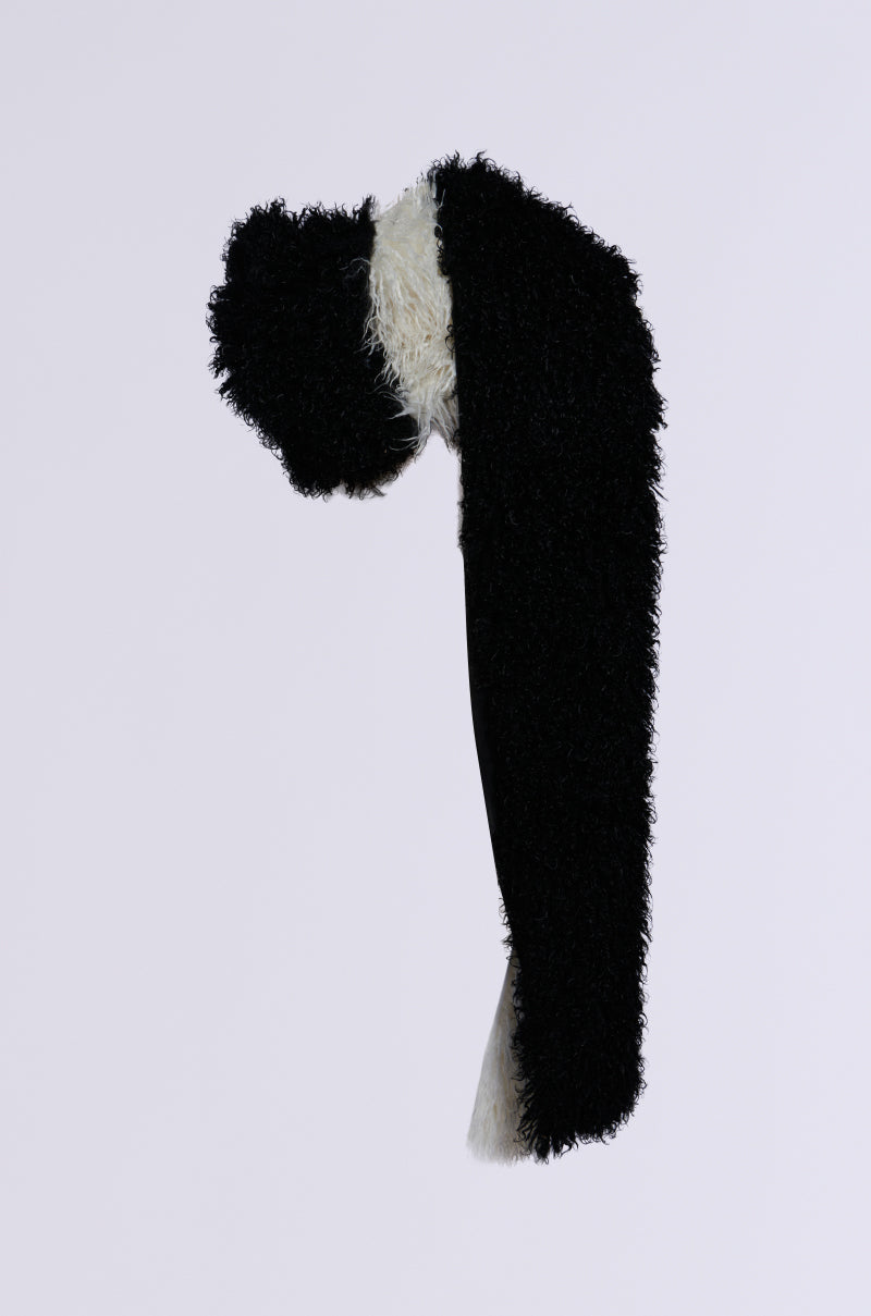 The EPIC DREAMS FAUX FUR SCARF, featuring a color block pattern in black and white with a polyester lining, hangs against a plain, light background. Its soft and textured appearance makes it a cozy and stylish winter accessory.