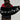 A person with long, wavy hair holds the F*CK IT FAUX FUR SCARF, a black faux fur shawl accented with white letters spelling "FAKE AS F*CK." They are dressed in a red top and a black lace skirt that offers an oversized fit, all set against a plain background.
