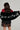 A person with long, wavy hair holds the F*CK IT FAUX FUR SCARF, a black faux fur shawl accented with white letters spelling "FAKE AS F*CK." They are dressed in a red top and a black lace skirt that offers an oversized fit, all set against a plain background.