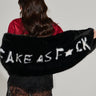 A person with long, wavy hair holds the F*CK IT FAUX FUR SCARF, a black faux fur shawl accented with white letters spelling "FAKE AS F*CK." They are dressed in a red top and a black lace skirt that offers an oversized fit, all set against a plain background.