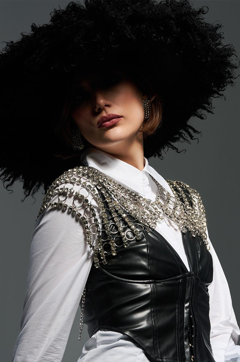 A woman poses against a gray background wearing the ALWAYS ELITE FAUX FUR BRIM HAT and an embellished vest over a white shirt. The vest features intricate silver detailing, adding a bold statement accessory to the look. Her expression is confident and poised.