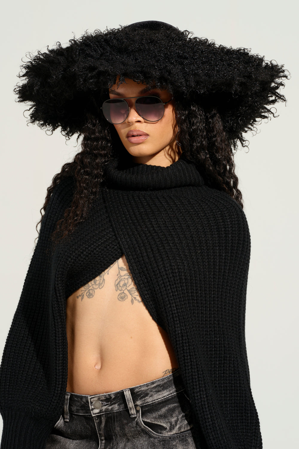 A person with long, curly hair and tattoos on their midriff pairs the ALWAYS ELITE FAUX FUR BRIM HAT with a pair of sunglasses and a black knit top. They complete their look with dark jeans against a plain, light backdrop.