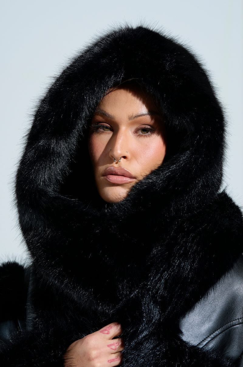 A person with a neutral expression is enveloped in the IM THE ONE FAUX FUR HOOD, covering their head and shoulders. The softly lit background highlights the texture of this winter accessory. 1