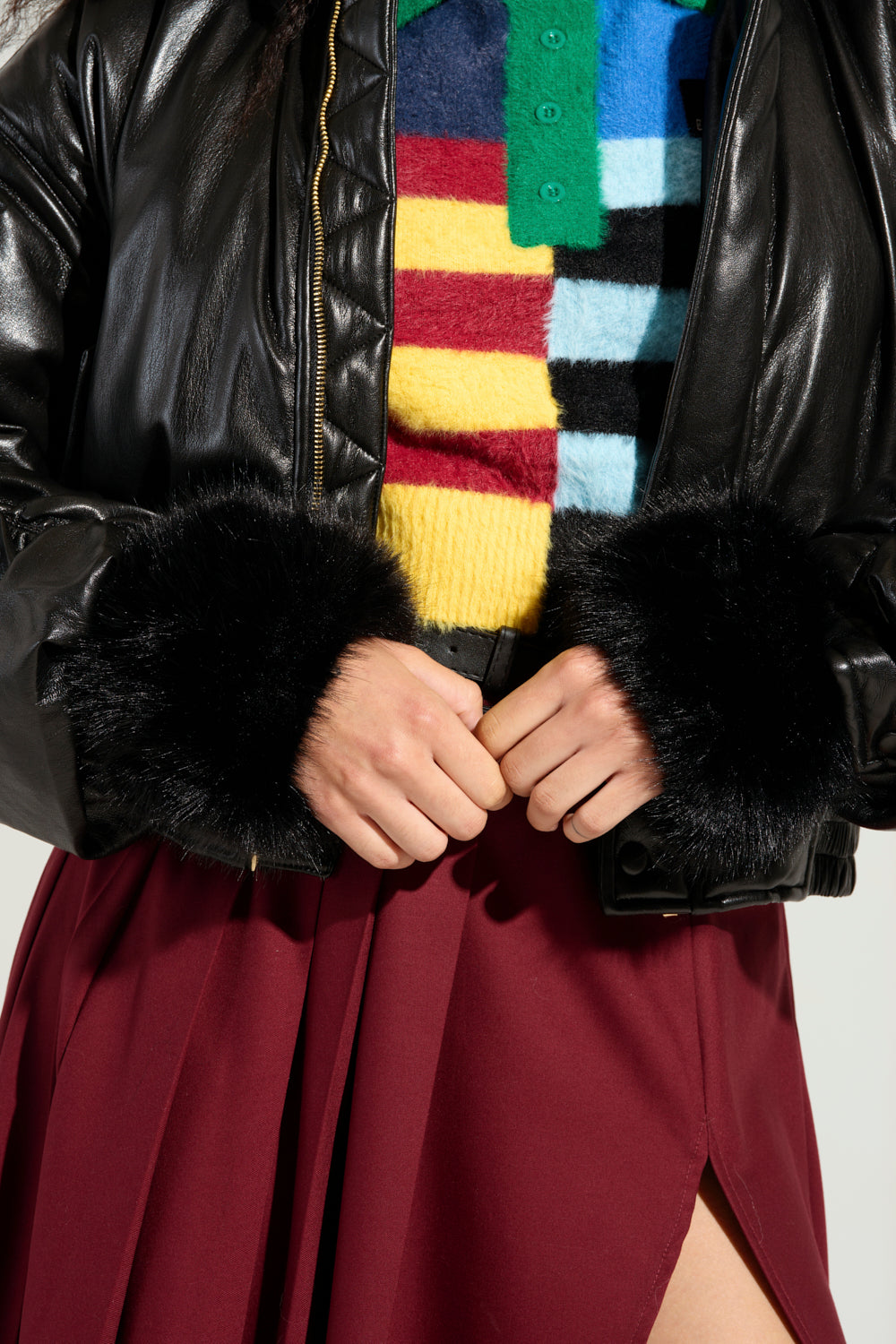 A person is styled in the STAYING WARM FAUX FUR CUFFS black leather jacket, layered over a vibrant striped sweater. They pair it with a burgundy skirt featuring a front slit as they gracefully pose against a plain background. Their hands are adjusting the jacket, emphasizing its customizable fit, ideal for complementing any winter ensemble.