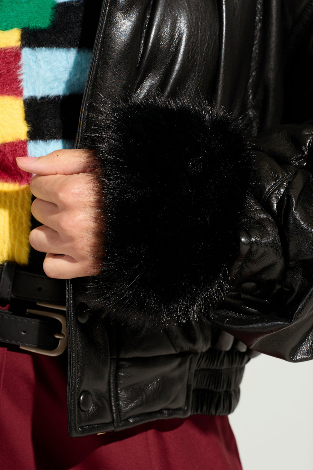 A person is shown wearing a colorful, checkered sweater under a black leather jacket featuring the STAYING WARM FAUX FUR CUFFS. The outfit is completed with maroon pants and a noticeable black belt, making it an ideal stylish winter accessory.