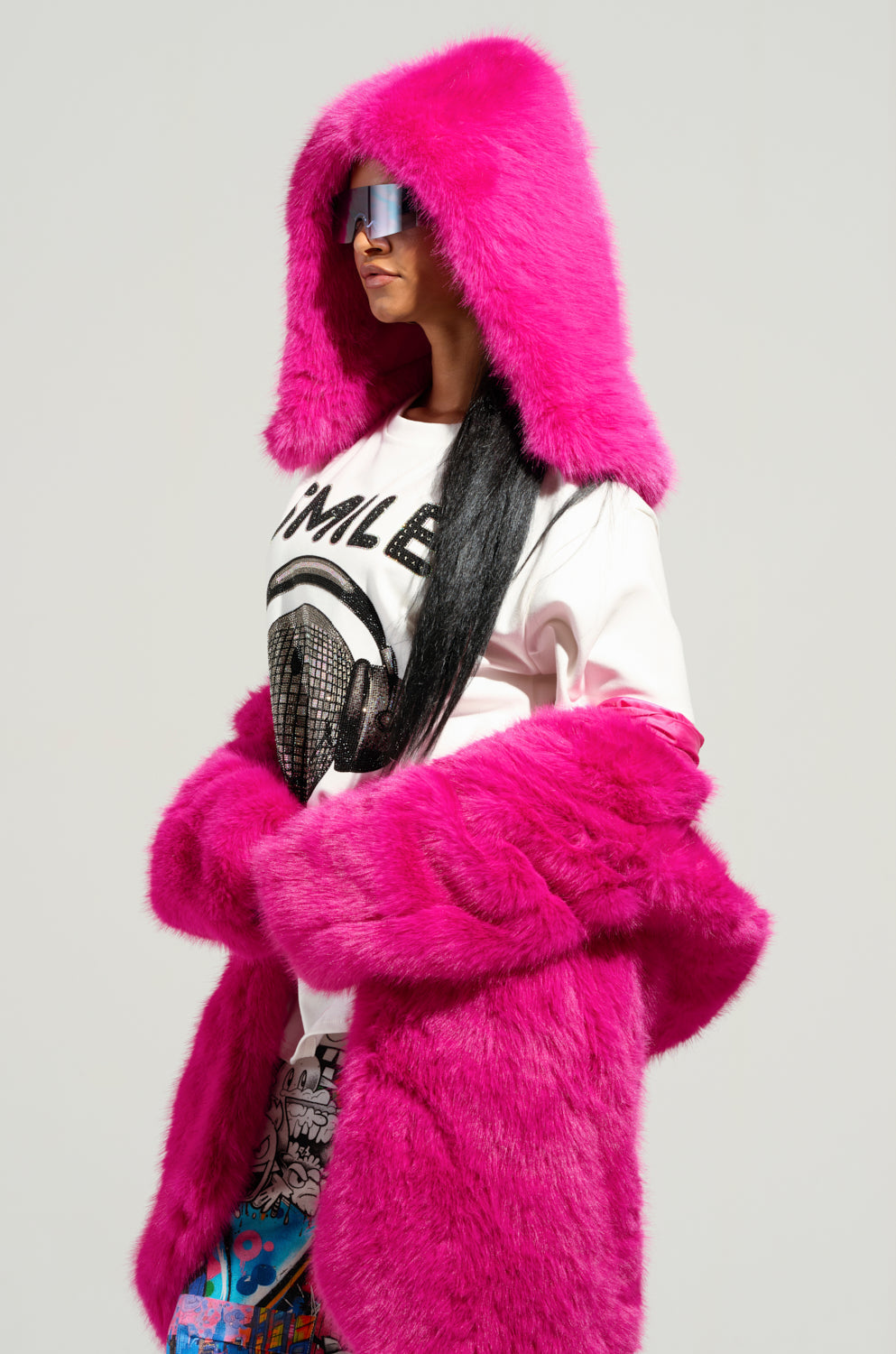 A person wears the STAND ON IT FAUX FUR HOOD in bright pink, paired with reflective sunglasses. Their long dark hair cascades over a white t-shirt featuring a graphic design, and colorful patterned pants add an extra flair. A plain, light-colored background completes this stylish look. 1