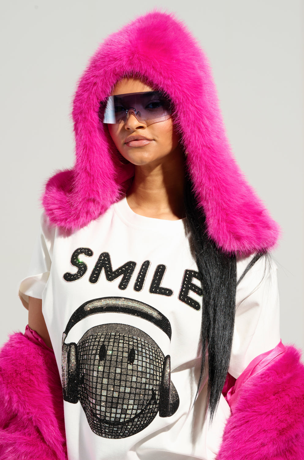 A person sporting a vibrant pink STAND ON IT FAUX FUR HOOD and matching gloves, complete with oversized sunglasses. They're wearing a white T-shirt featuring a graphic of headphones and "SMILE" boldly written in black letters. The plain gray backdrop highlights their bold style. 2