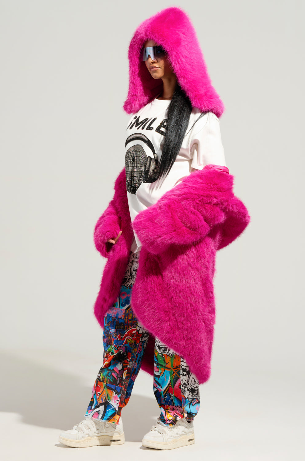 The individual is wearing a STAND ON IT FAUX FUR HOOD in vibrant pink, paired with mirrored sunglasses and a white graphic t-shirt. They complement their look with printed colorful pants and slip-on fit white sneakers, all against a plain background. 3
