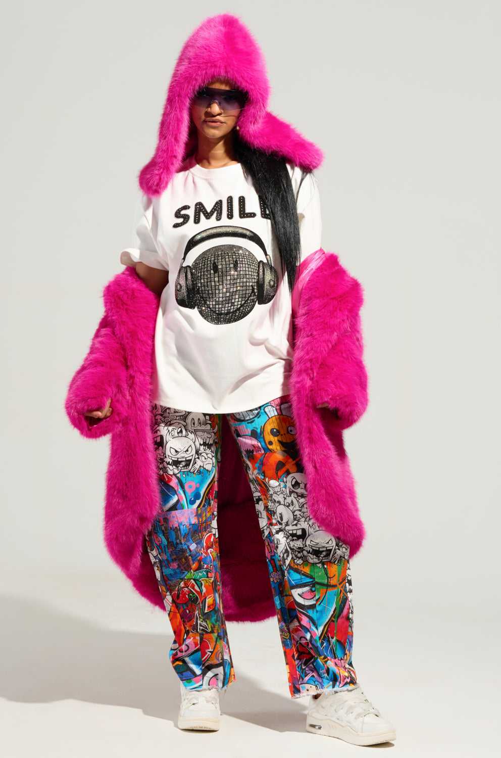 Wearing a STAND ON IT FAUX FUR HOOD in a vivid pink shade over a white "SMILE" t-shirt featuring a disco ball design, the individual confidently complements their outfit with vibrant, graphic-patterned pants and white slip-on fit sneakers, presenting a striking silhouette against the plain background. 4