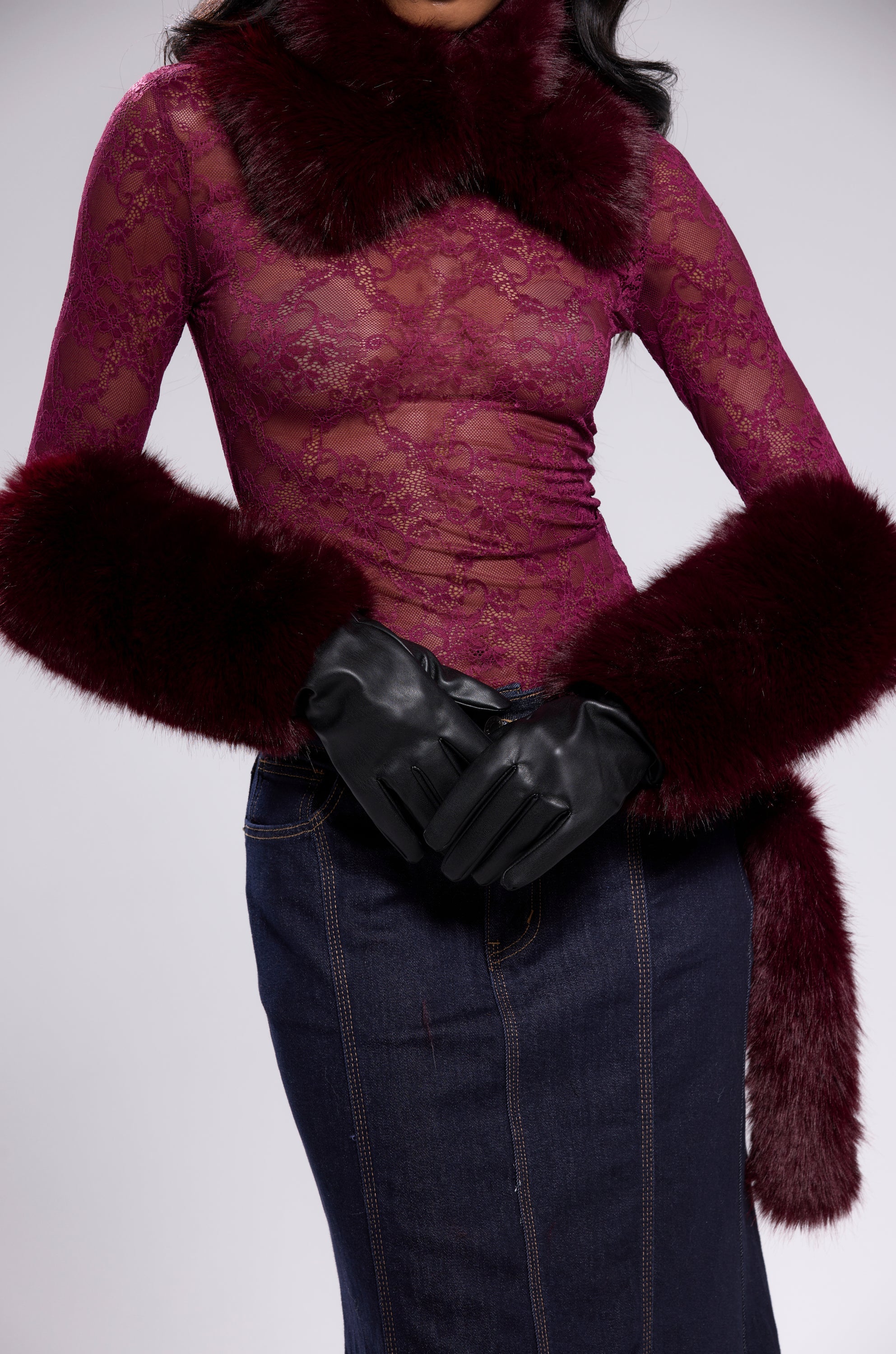 A person wears a sheer burgundy lace top with faux fur trim, a matching fur collar, ALAIA FAUX FUR GLOVES, and a dark denim skirt. The head is partially out of frame against a plain, light background. 0