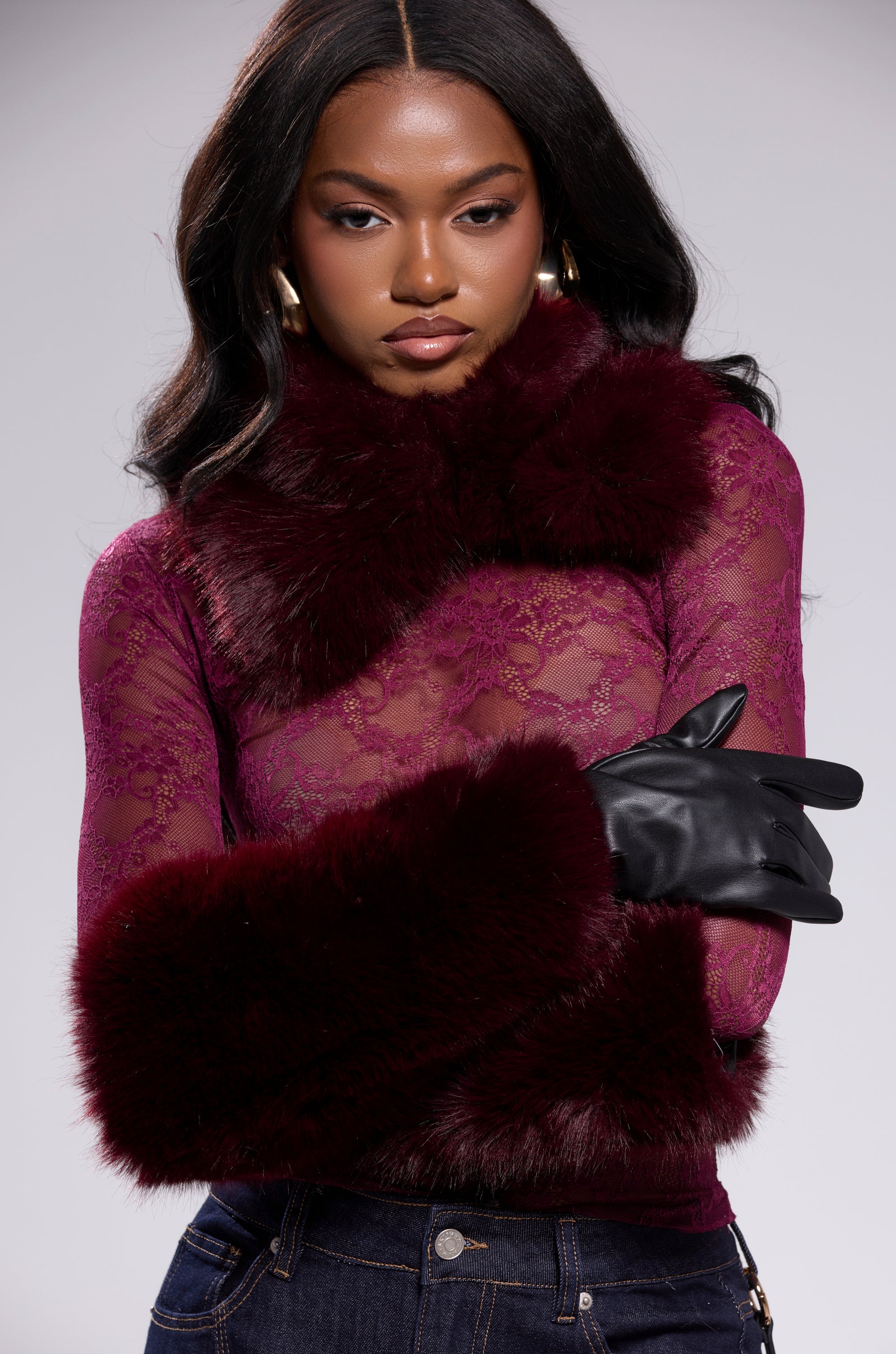 A woman wearing plum ALAIA FAUX FUR GLOVES, a sheer burgundy lace top with faux fur collar and cuffs, gold hoop earrings, and dark jeans poses with arms crossed against a neutral background. 1