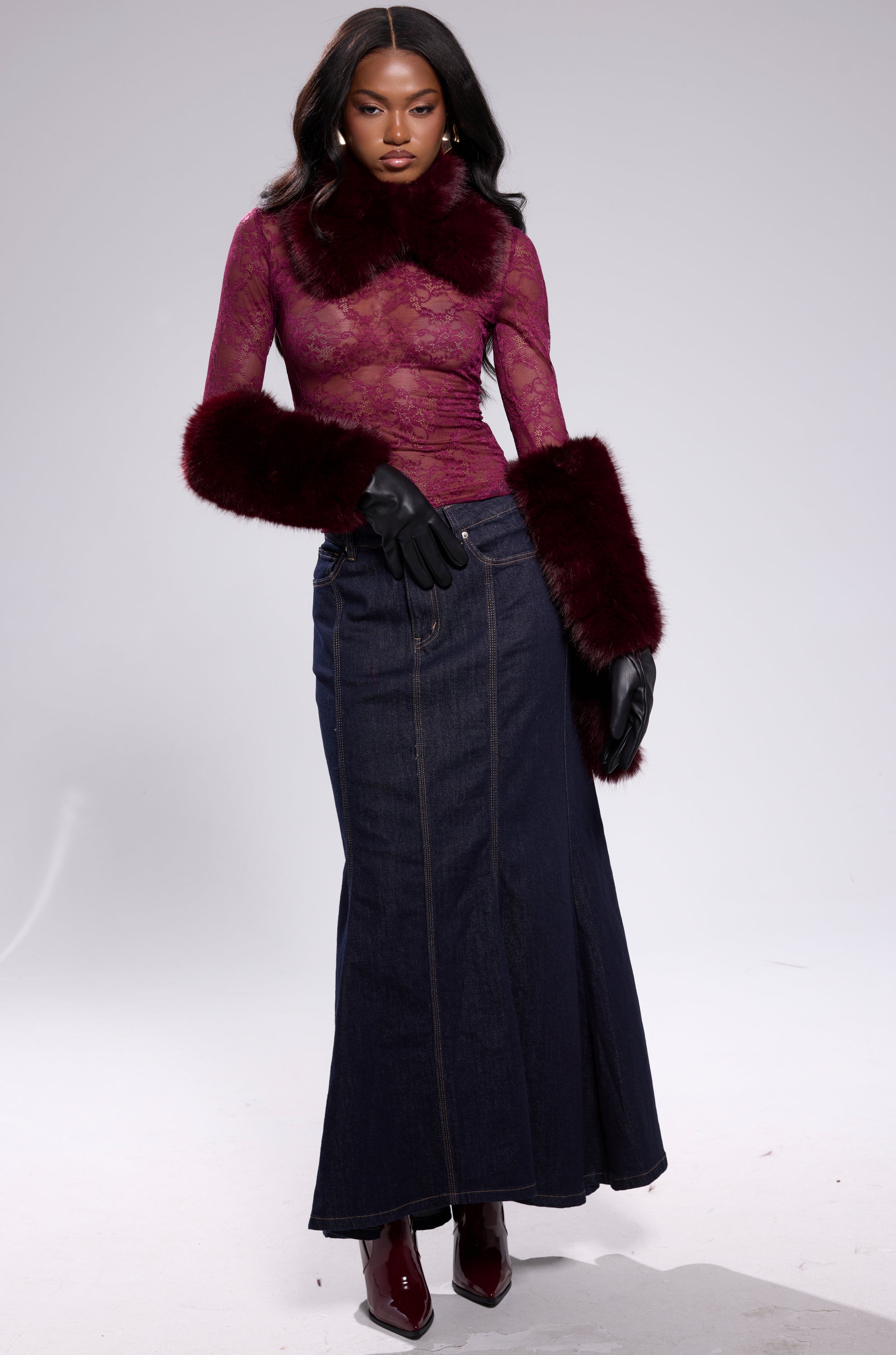 A woman poses in elbow-length ALAIA FAUX FUR GLOVES, a burgundy lace top with matching fur cuffs and collar, a dark denim skirt, and burgundy boots against a plain light gray background. 5