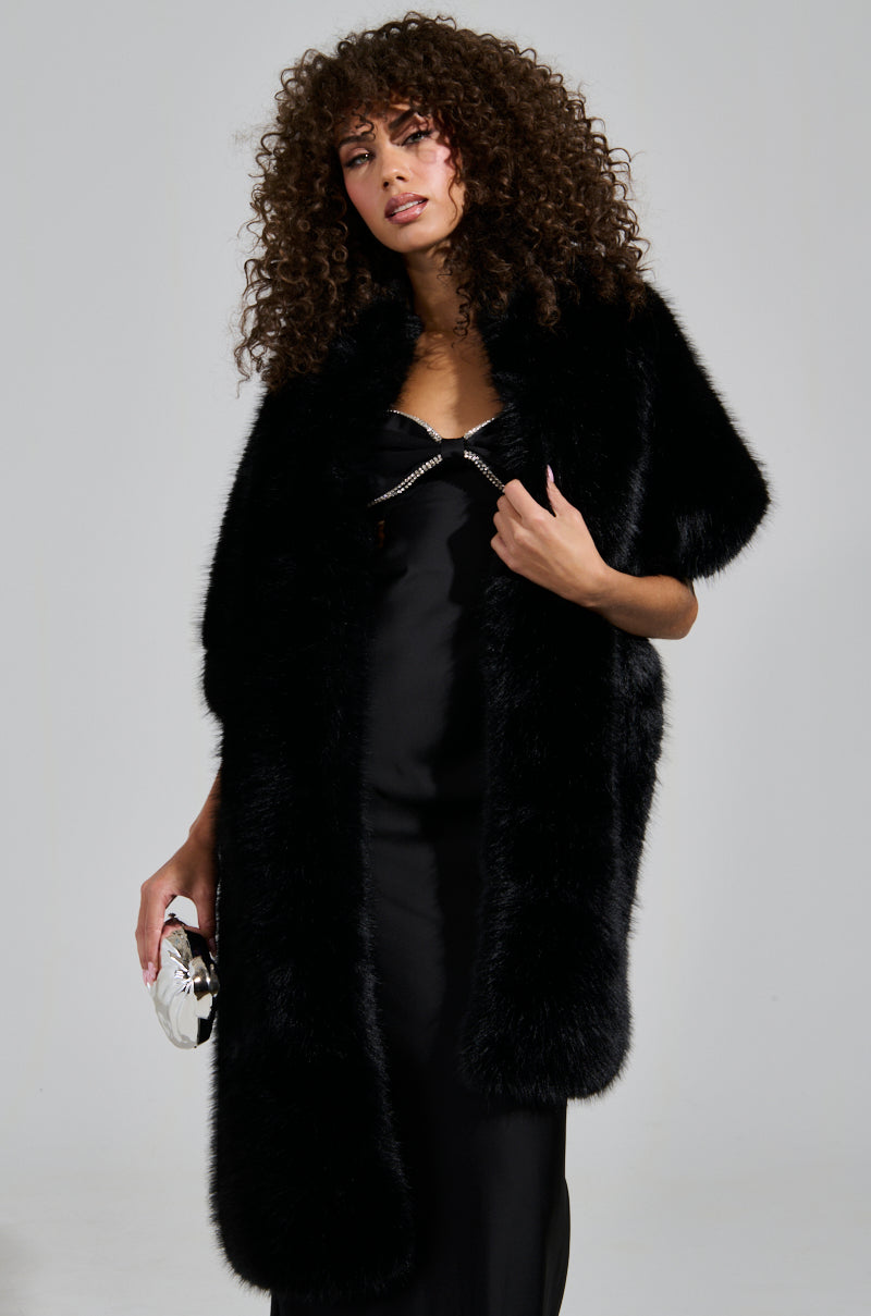 A person with curly hair wears the SOFT LIFE FAUX FUR SCARF draped elegantly over a black evening gown, clutching a small round handbag. They stand against a plain background, exuding elegance and sophistication.
