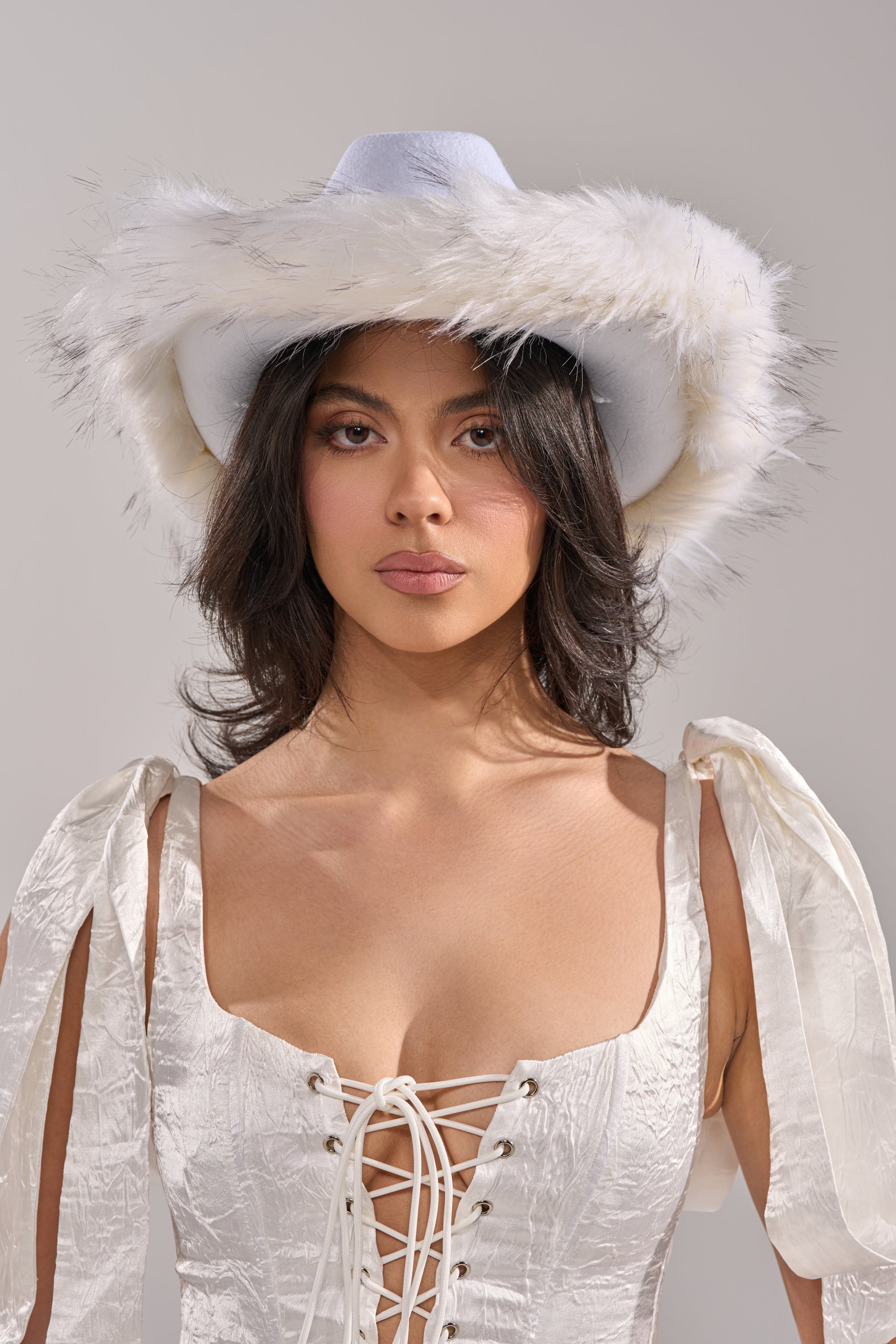 A woman in a white satin lace-up dress and the LETS RIDE FAUX FUR TRIM COWBOY HAT stands against a plain light gray background. 2