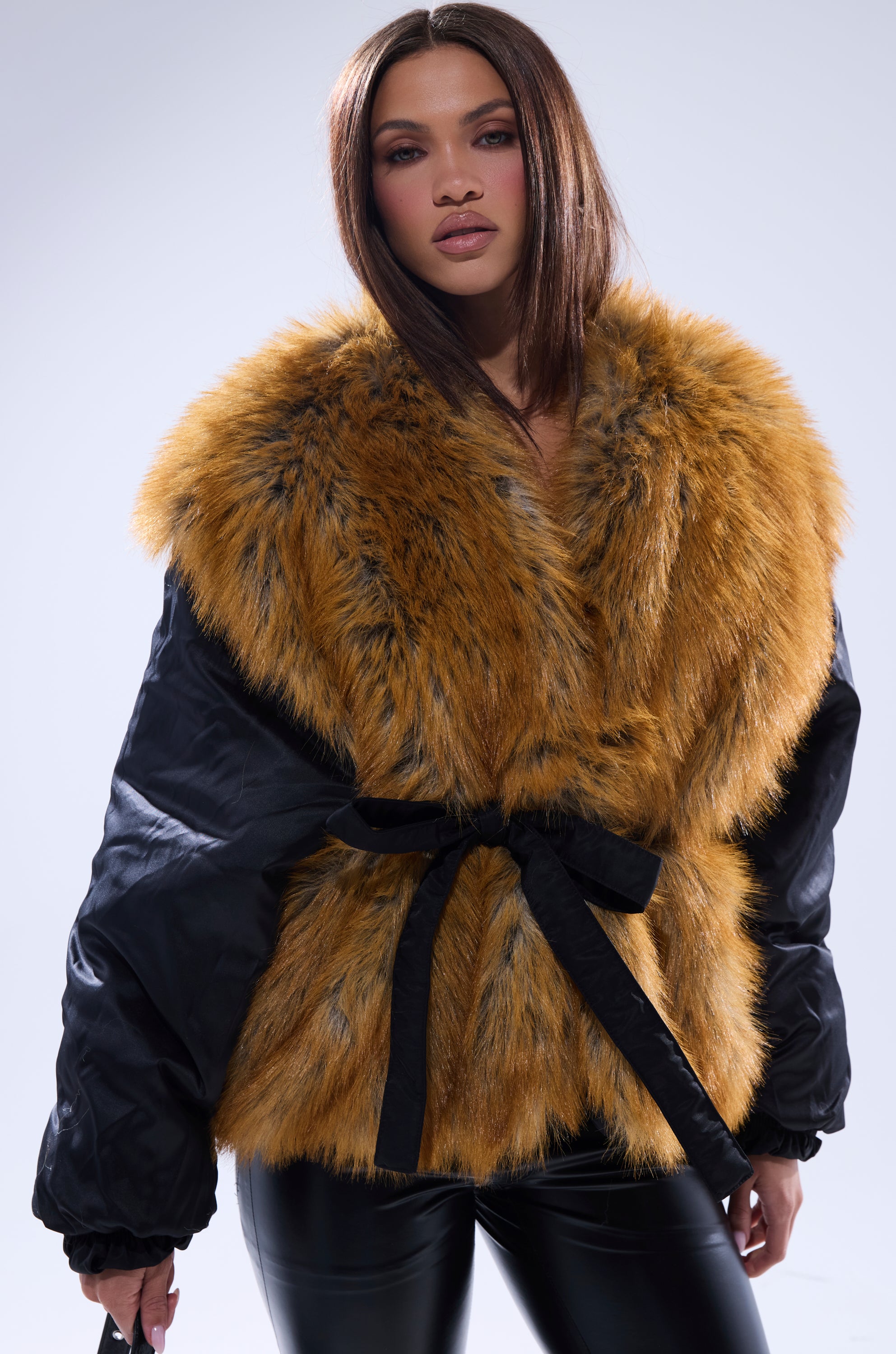 A woman with straight brown hair models the INSIDE OUT REVERSIBLE PUFFER, a large golden-brown faux fur jacket with black sleeves and belt, paired with black leather pants against a plain light background, adding chic warmth to her outfit.