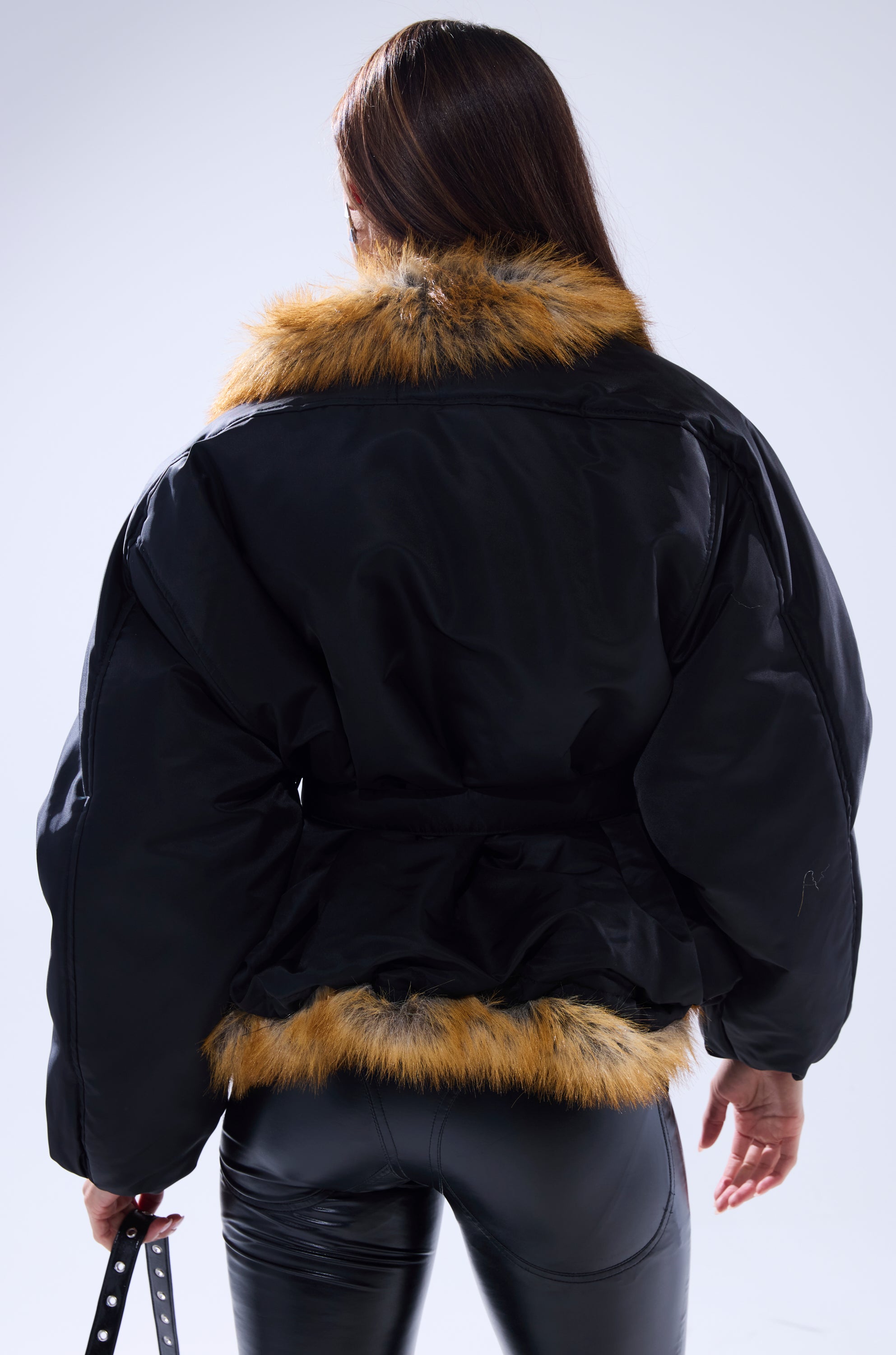 A woman with long brown hair, seen from behind, is wearing the INSIDE OUT REVERSIBLE PUFFER with brown collar and hem trim, paired with black leather pants. She holds a strap in her left hand.
