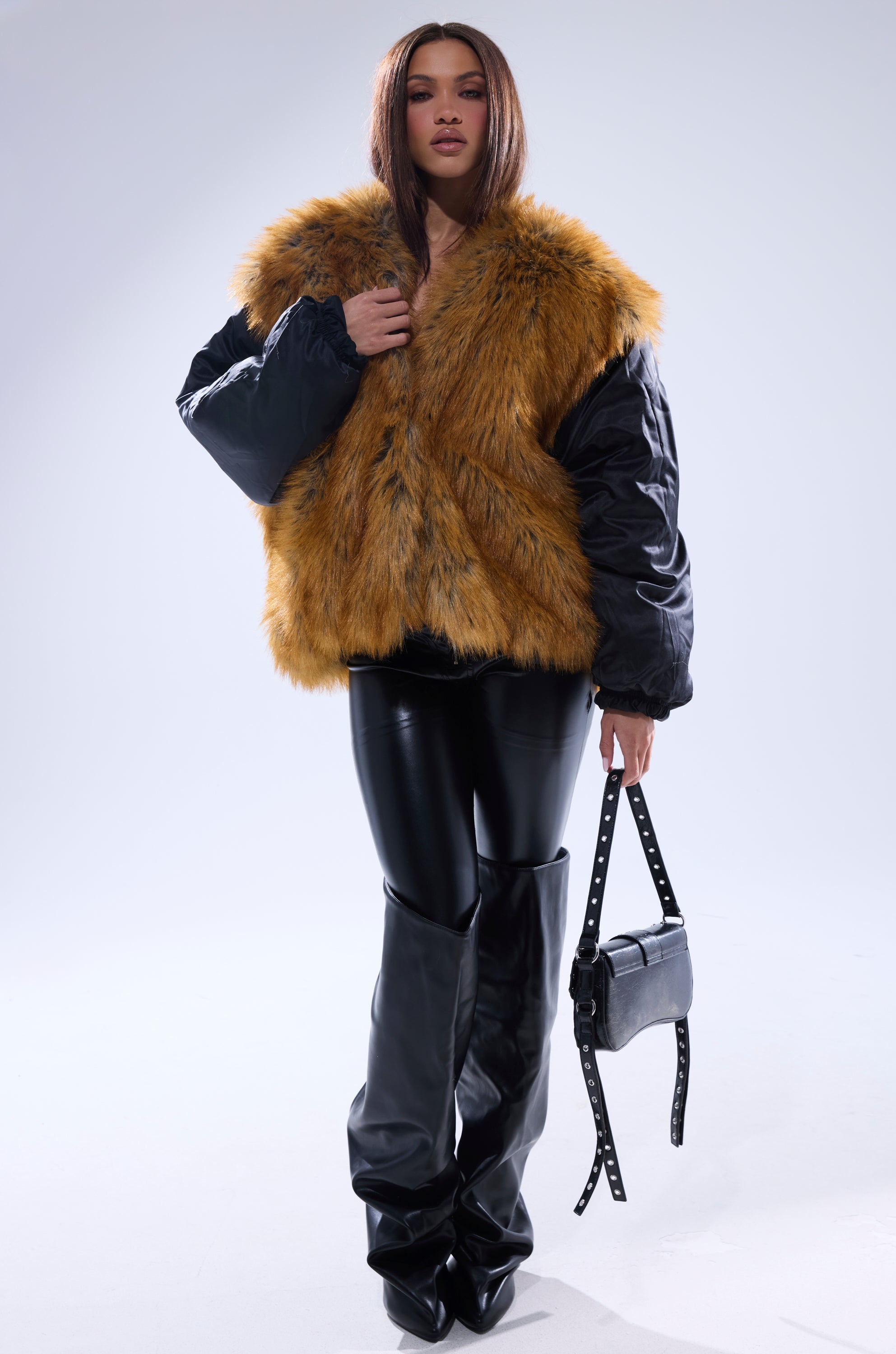 A woman stands confidently in the INSIDE OUT REVERSIBLE PUFFER, paired with black leather pants, knee-high boots, and a black studded handbag against a light background.