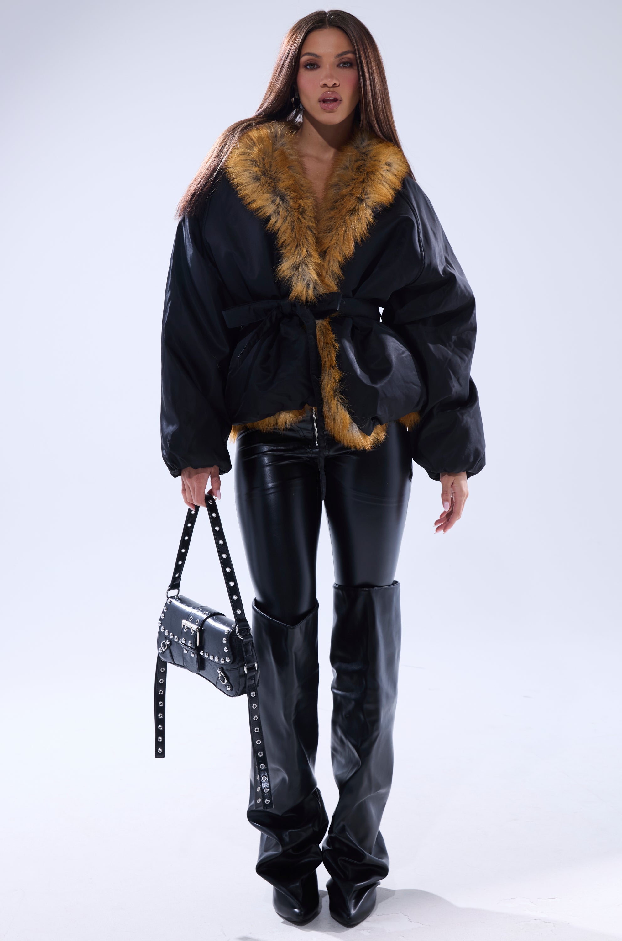 A woman models the INSIDE OUT REVERSIBLE PUFFER, styled with black leather pants, tall black boots, a studded handbag, and bold makeup. She stands against a plain light background with long hair.