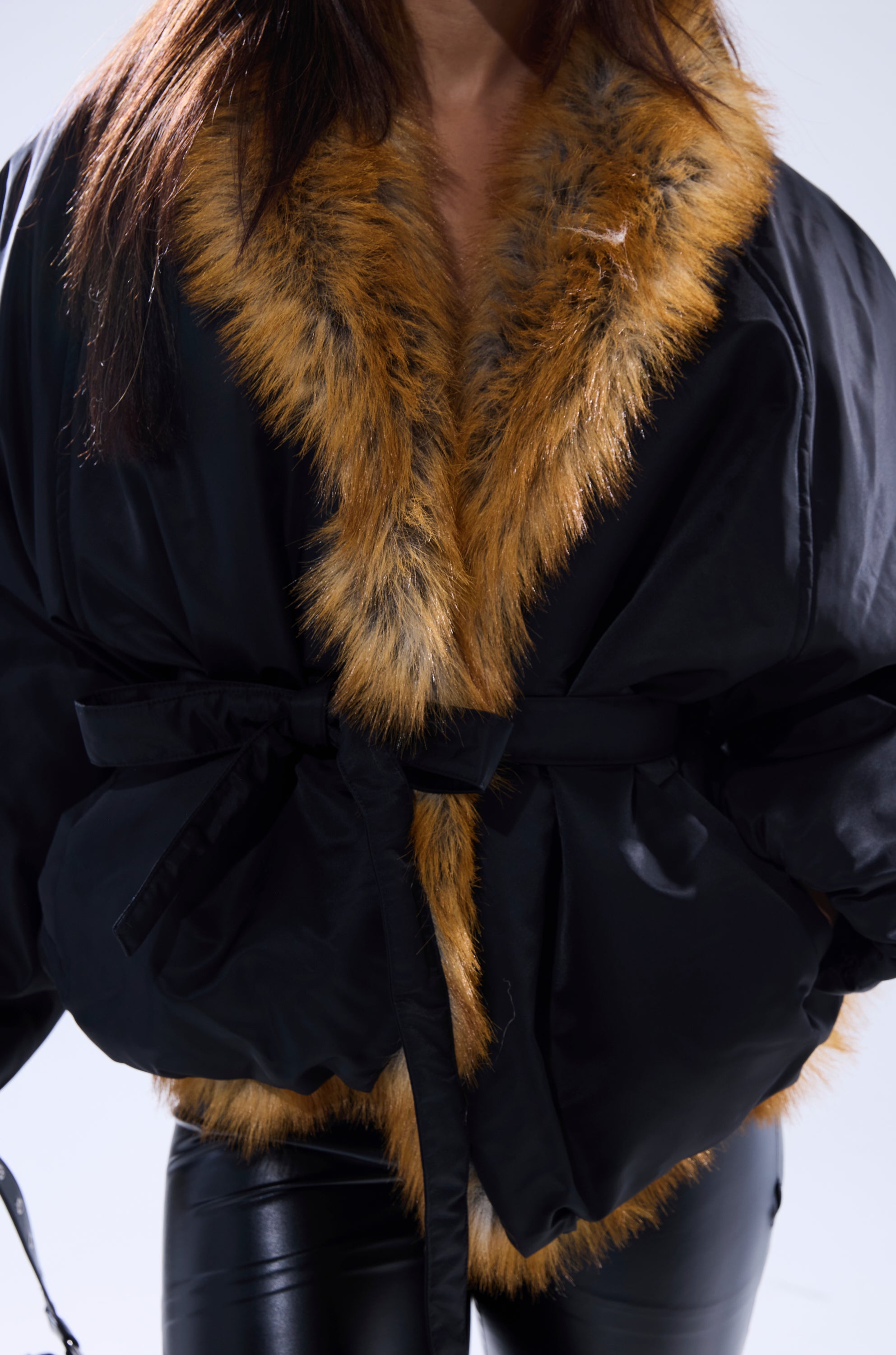 A person wears the INSIDE OUT REVERSIBLE PUFFER with a wide, fluffy brown collar, tied at the waist with a matching belt, paired with black leather pants. The background is plain and light-colored.