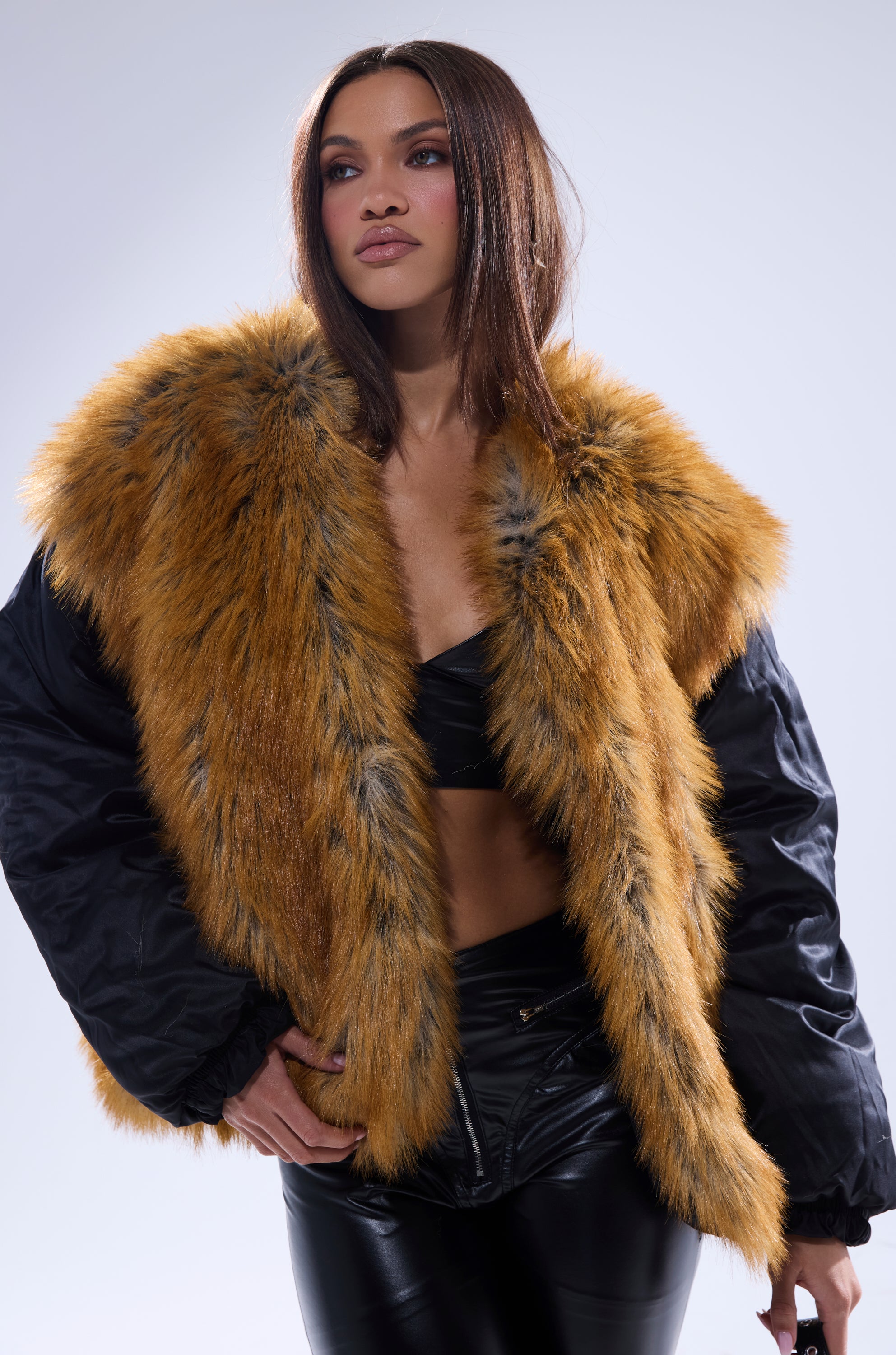 A woman poses confidently in the INSIDE OUT REVERSIBLE PUFFER, a stylish faux fur jacket with golden-brown and black tones, over a black crop top and leather pants against a light gray background.