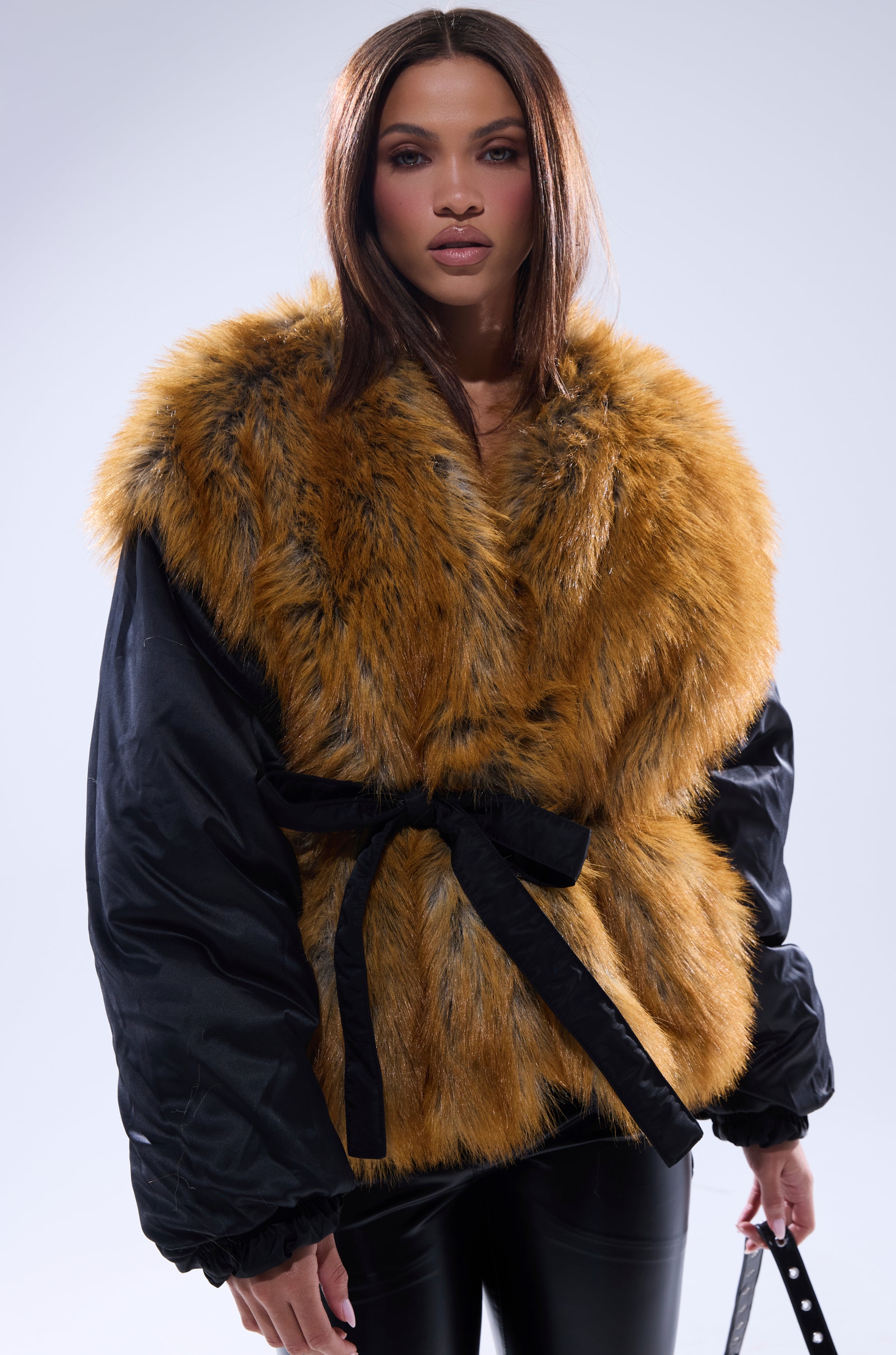 A woman poses in the INSIDE OUT REVERSIBLE PUFFER, a black faux fur jacket with oversized golden-brown trim and a black tie belt, styled with black leather pants against a plain light gray background.
