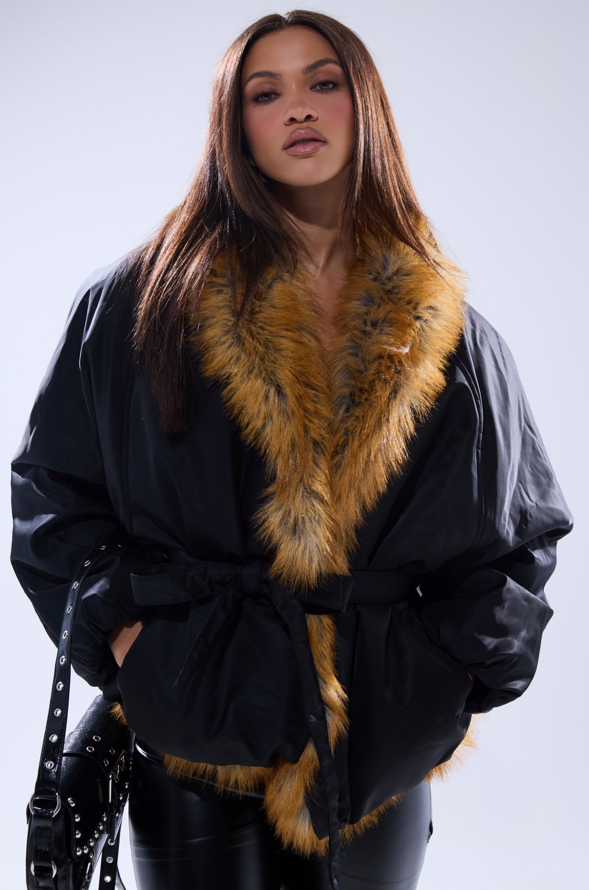 A woman with straight brown hair confidently models the INSIDE OUT REVERSIBLE PUFFER, featuring a black faux fur exterior with a wide brown collar and leather belt, styled with black leather pants against a plain light background.
