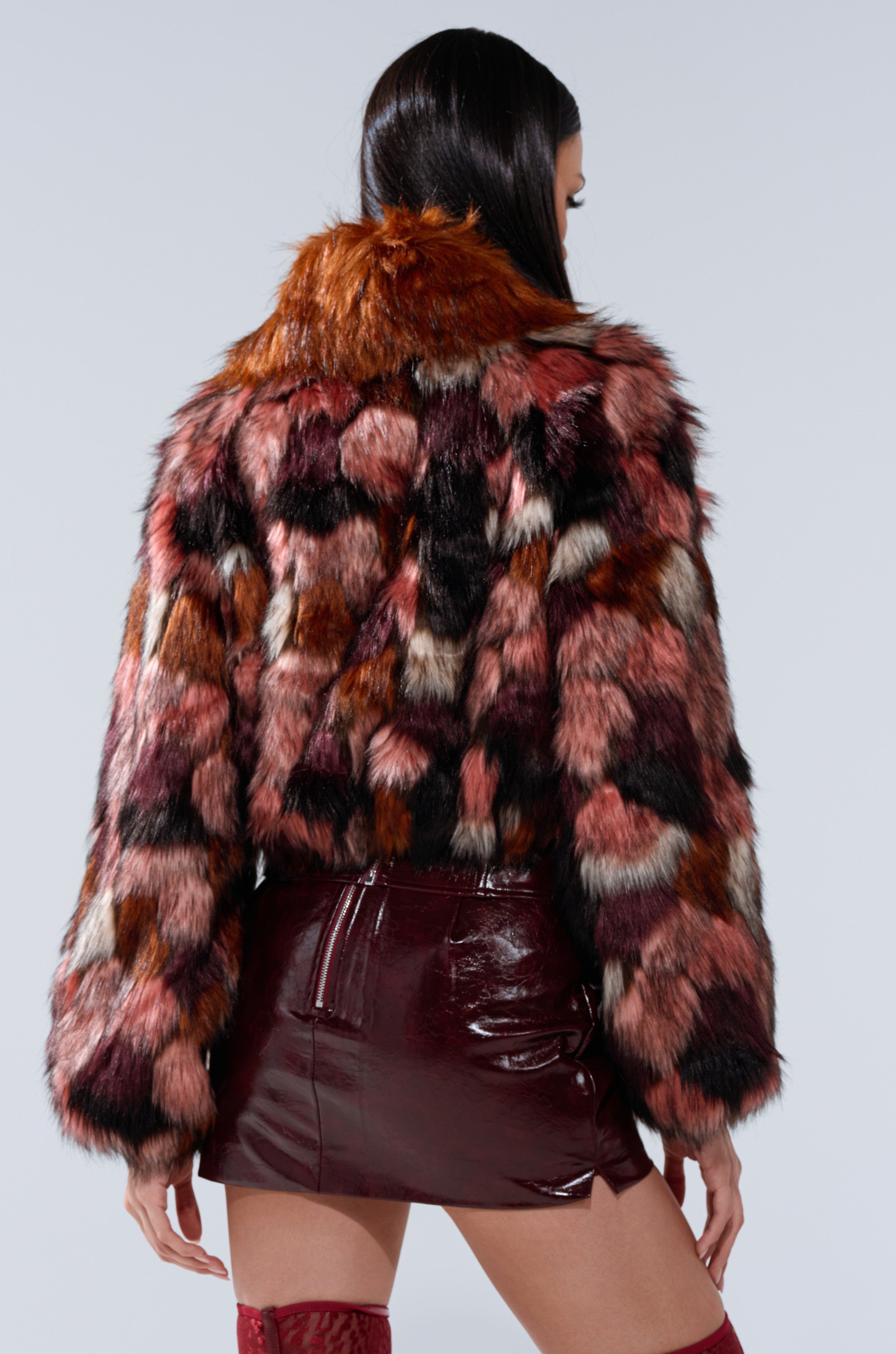 A woman with long dark hair, seen from the back, wears the DELTA FAUX FUR COAT in red, pink, black, and white over a shiny burgundy mini skirt and red thigh-high boots.