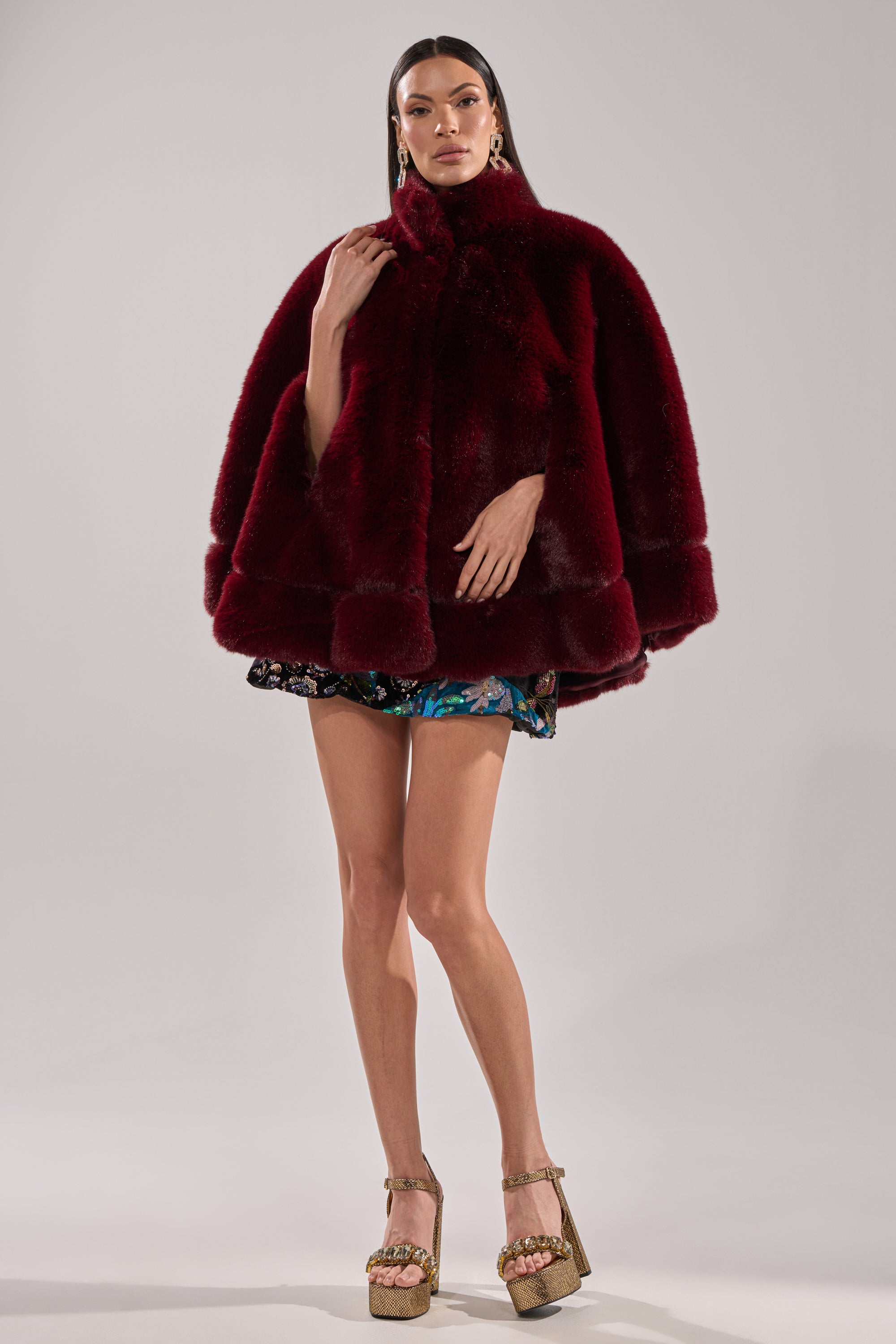 A woman poses confidently against a light gray background, wearing the CELINE FAUX FUR CAPE in deep burgundy, paired with a short floral skirt, gold platform heels, and large gold earrings.