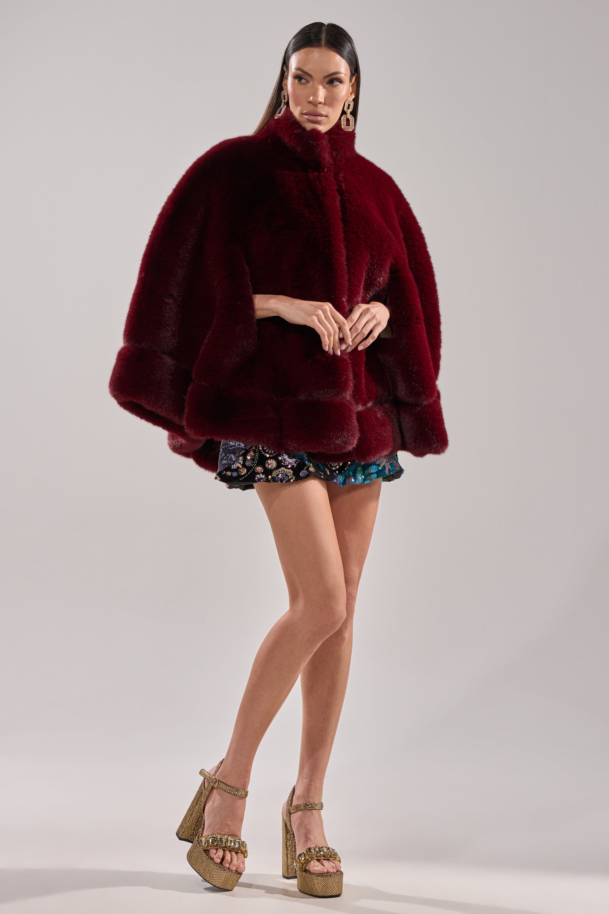 A woman models the CELINE FAUX FUR CAPE in deep red with short patterned shorts, gold platform heels, and statement earrings, posing against a simple light background.