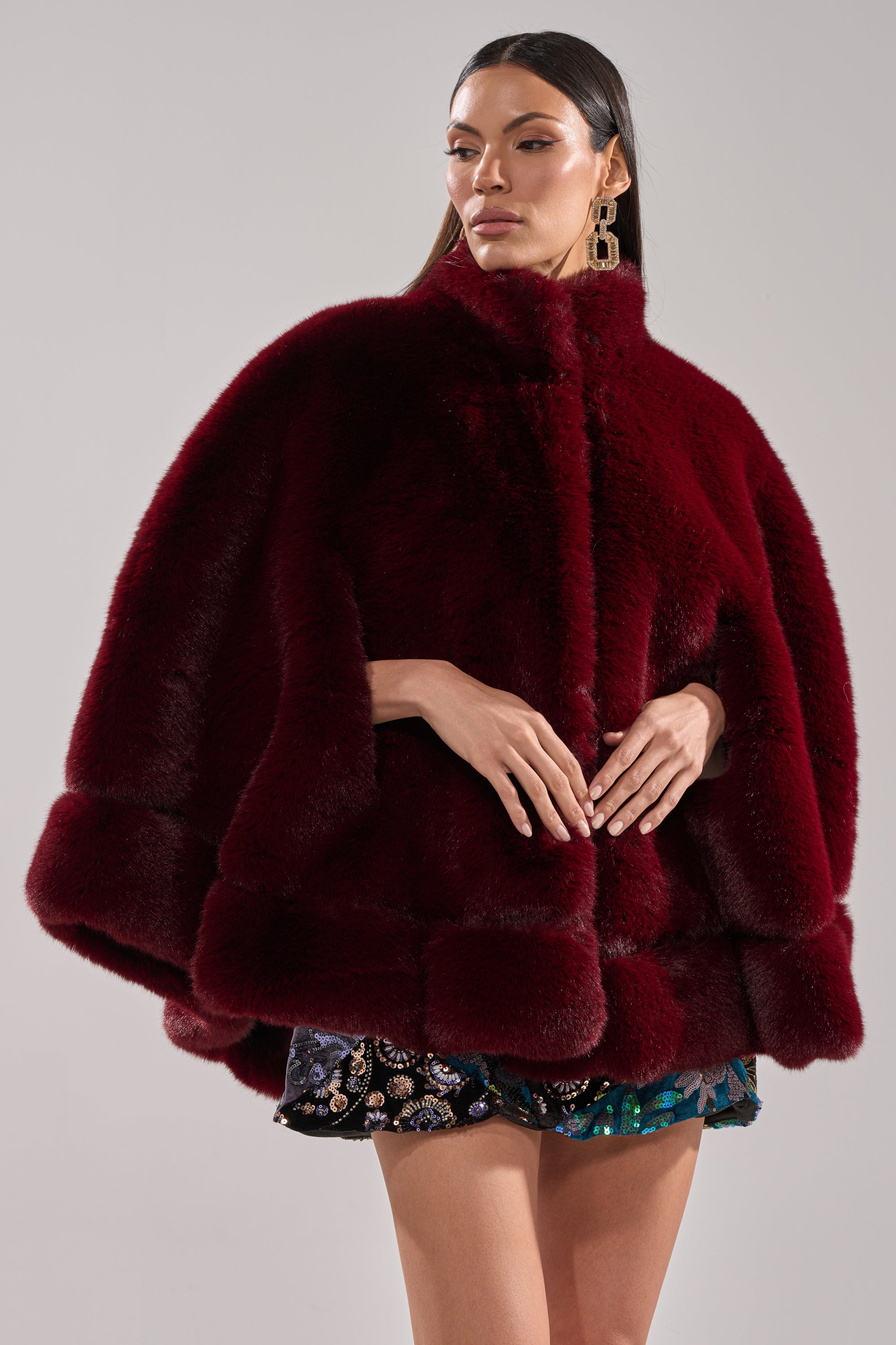 A woman with straight dark hair and gold statement earrings wears the luxurious deep red CELINE FAUX FUR CAPE over a short patterned skirt, standing with her hands clasped against a plain light gray background.