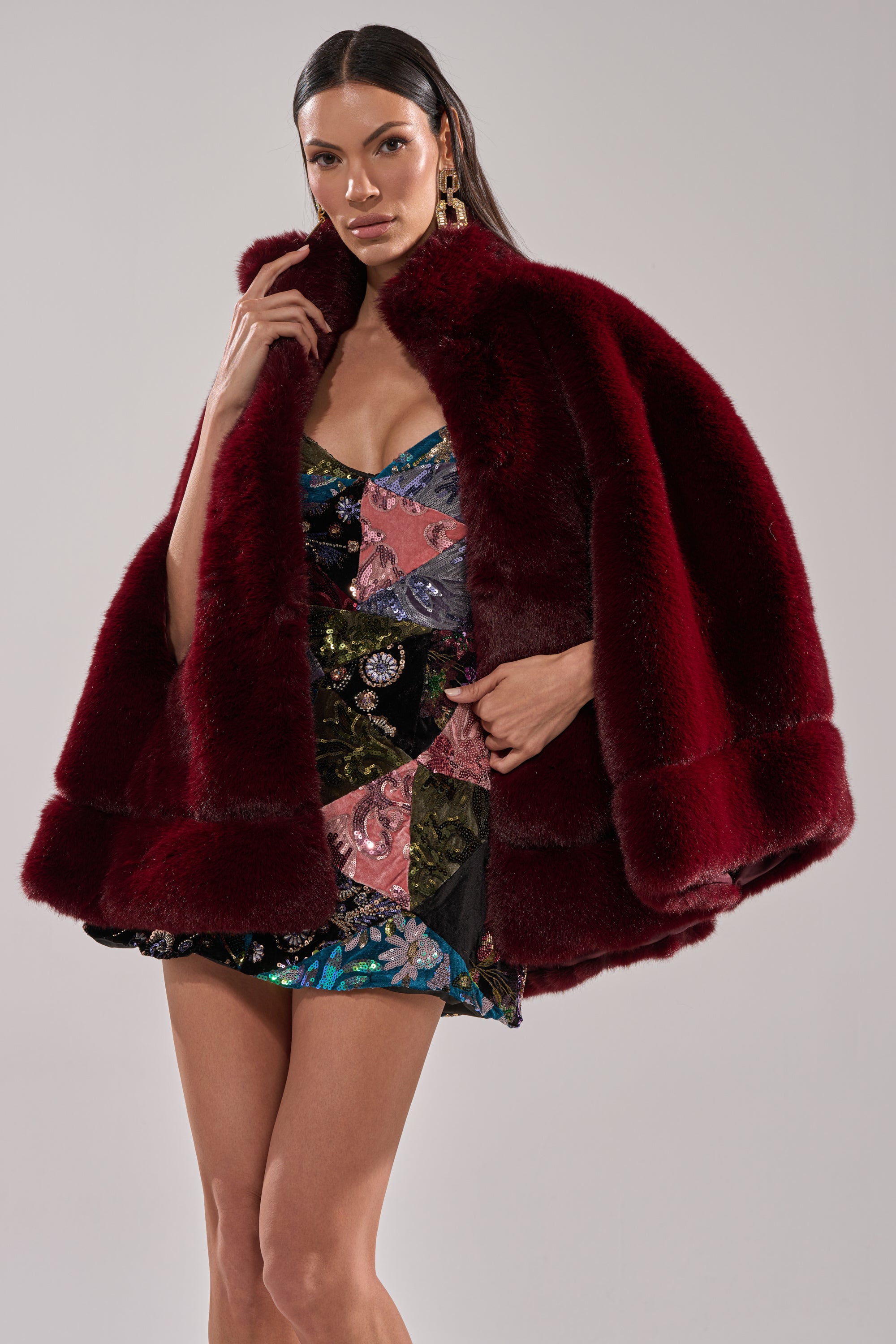 A woman with long dark hair and bold earrings poses in the CELINE FAUX FUR CAPE, a rich burgundy women's cape, layered over a colorful floral mini dress against a plain light gray background.