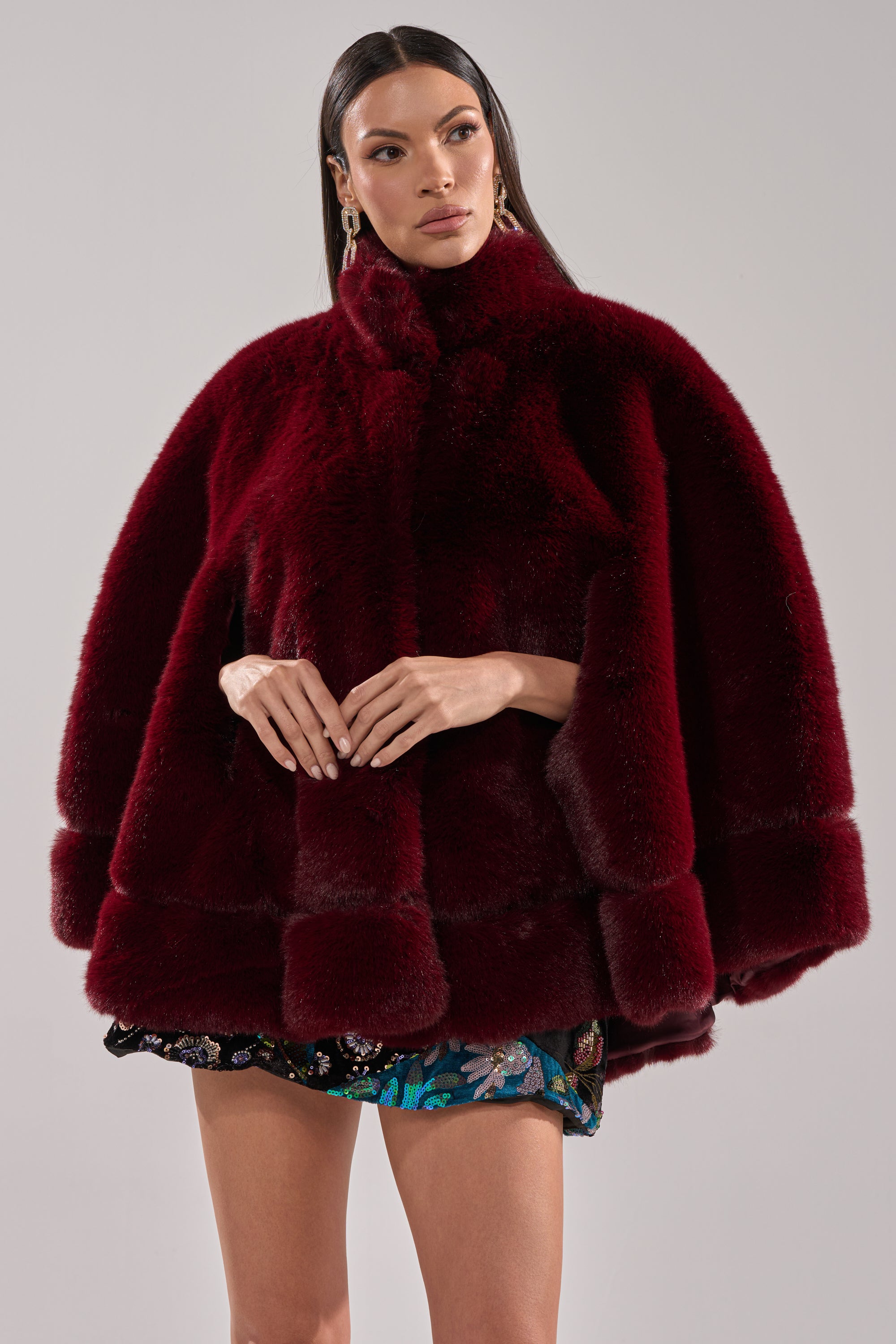 A woman with long dark hair and gold earrings poses against a plain light background wearing the CELINE FAUX FUR CAPE in deep burgundy with wide sleeves, paired with a short floral-patterned skirt.