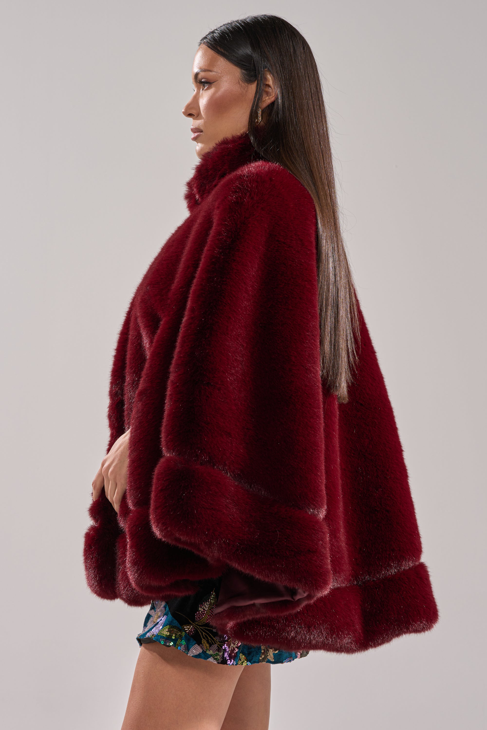 A woman with long straight hair in profile stands against a plain light background, wearing the CELINE FAUX FUR CAPE in deep red paired with patterned shorts.