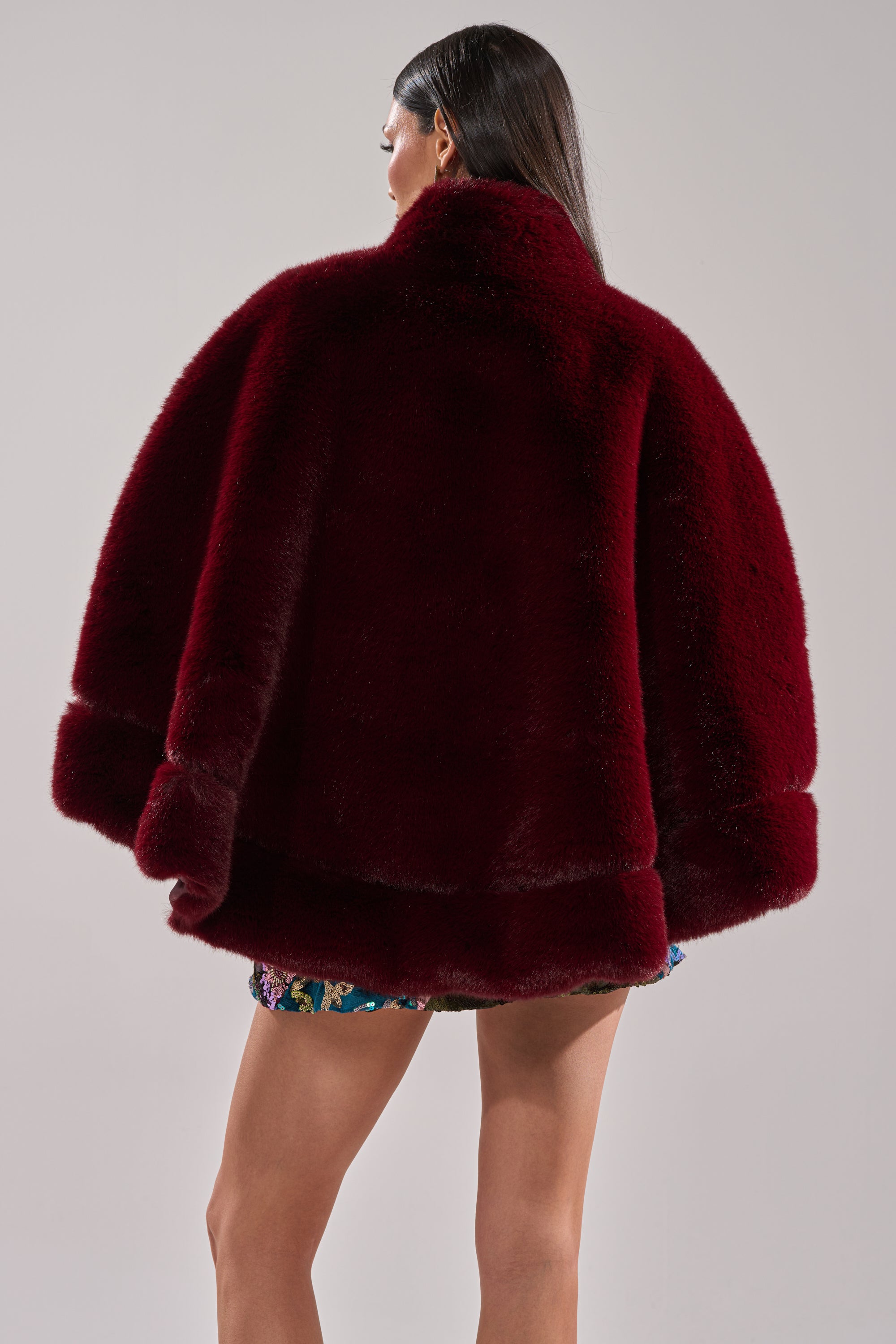 A person with long dark hair is seen from behind wearing the CELINE FAUX FUR CAPE with a mock neck and satin lining over a vibrant sequined skirt, set against a plain light background.