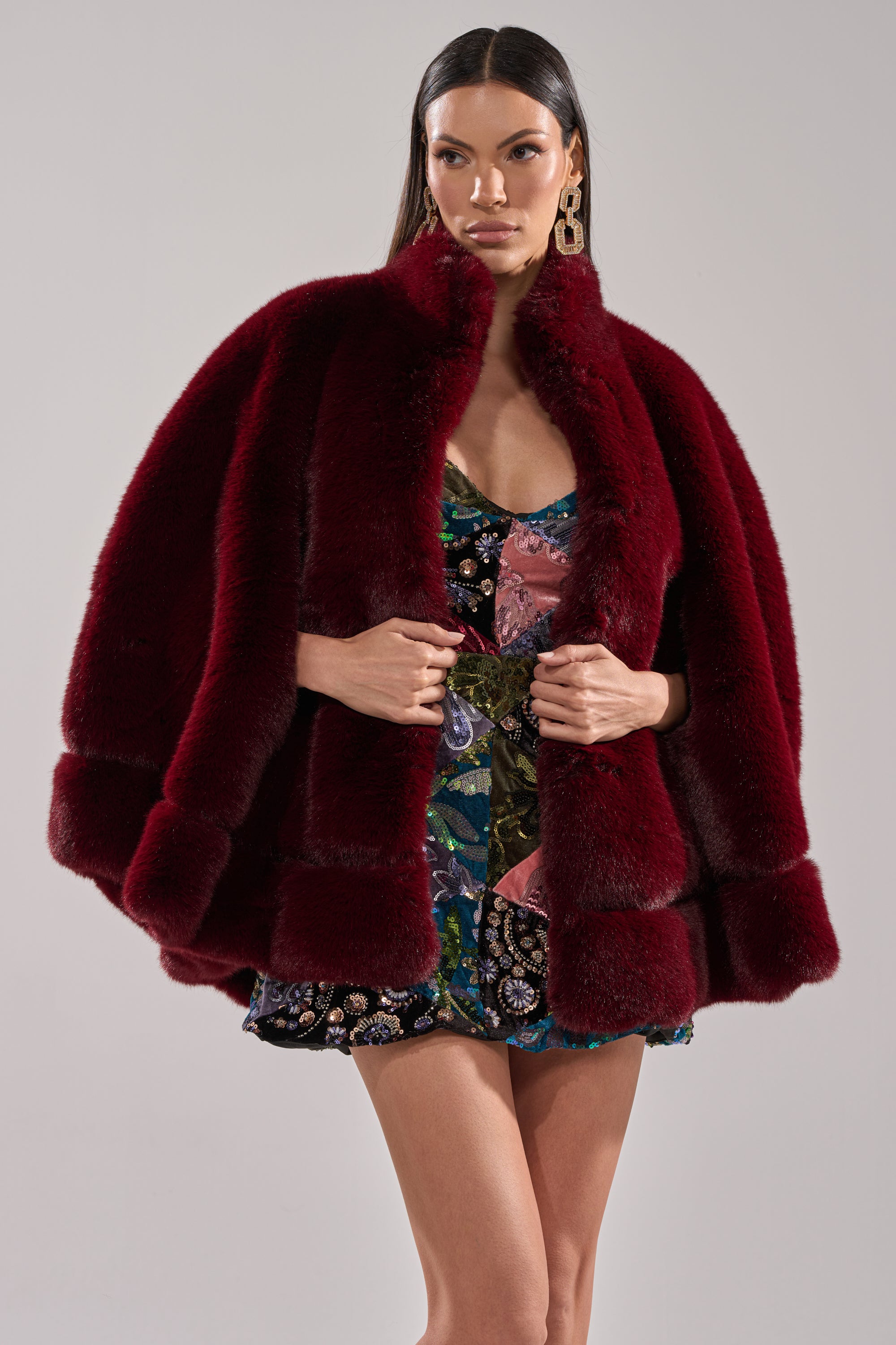 A woman poses against a light background, wearing the CELINE FAUX FUR CAPE with satin lining over a vibrant floral mini dress, her hands clasped at the front and large statement earrings completing the look.