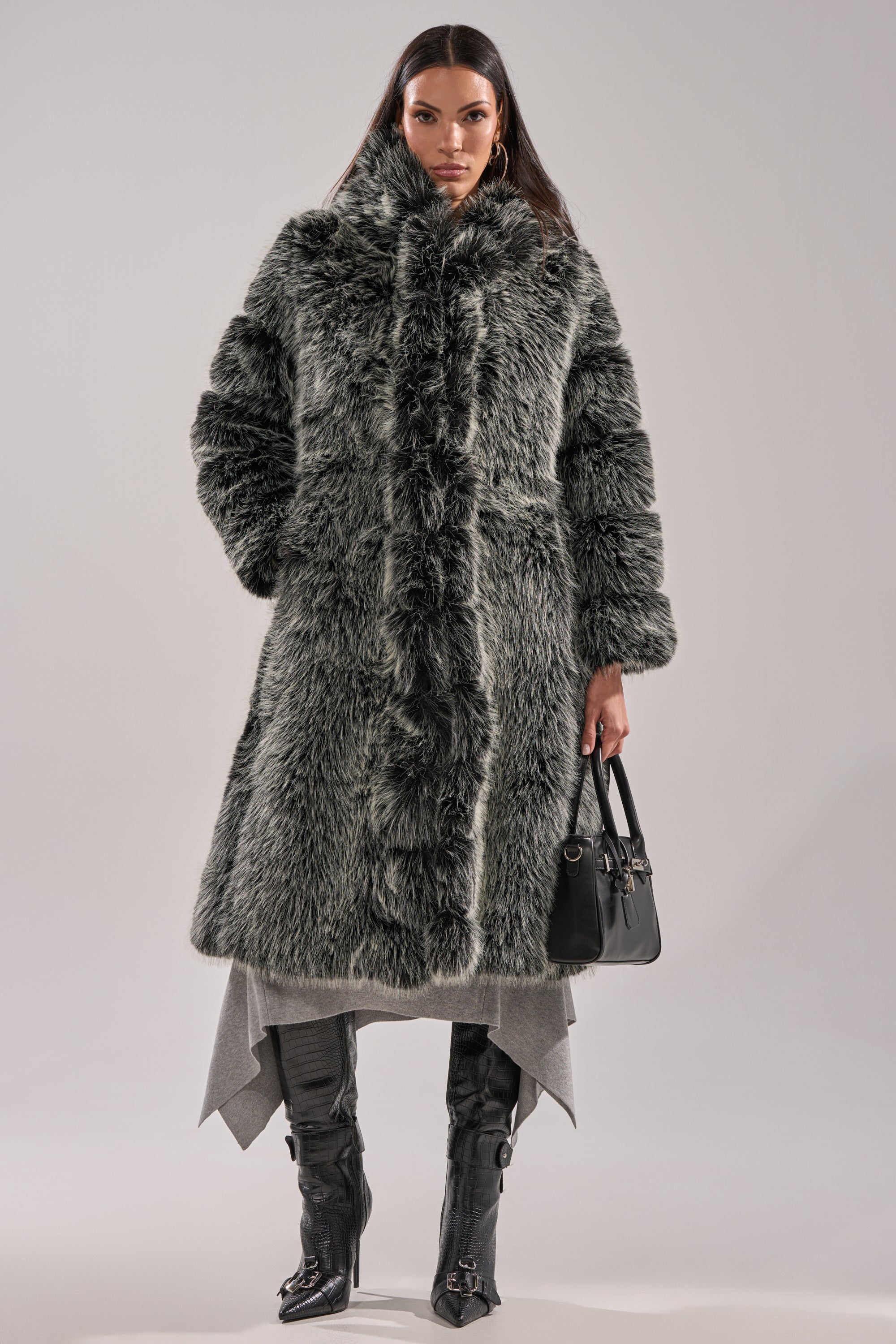 A woman stands facing forward against a plain light background, wearing the SNOW BUNNY HOODED FAUX FUR COAT—a chic longline gray faux fur coat—paired with a gray layered skirt, black knee-high boots, and holding a black handbag.