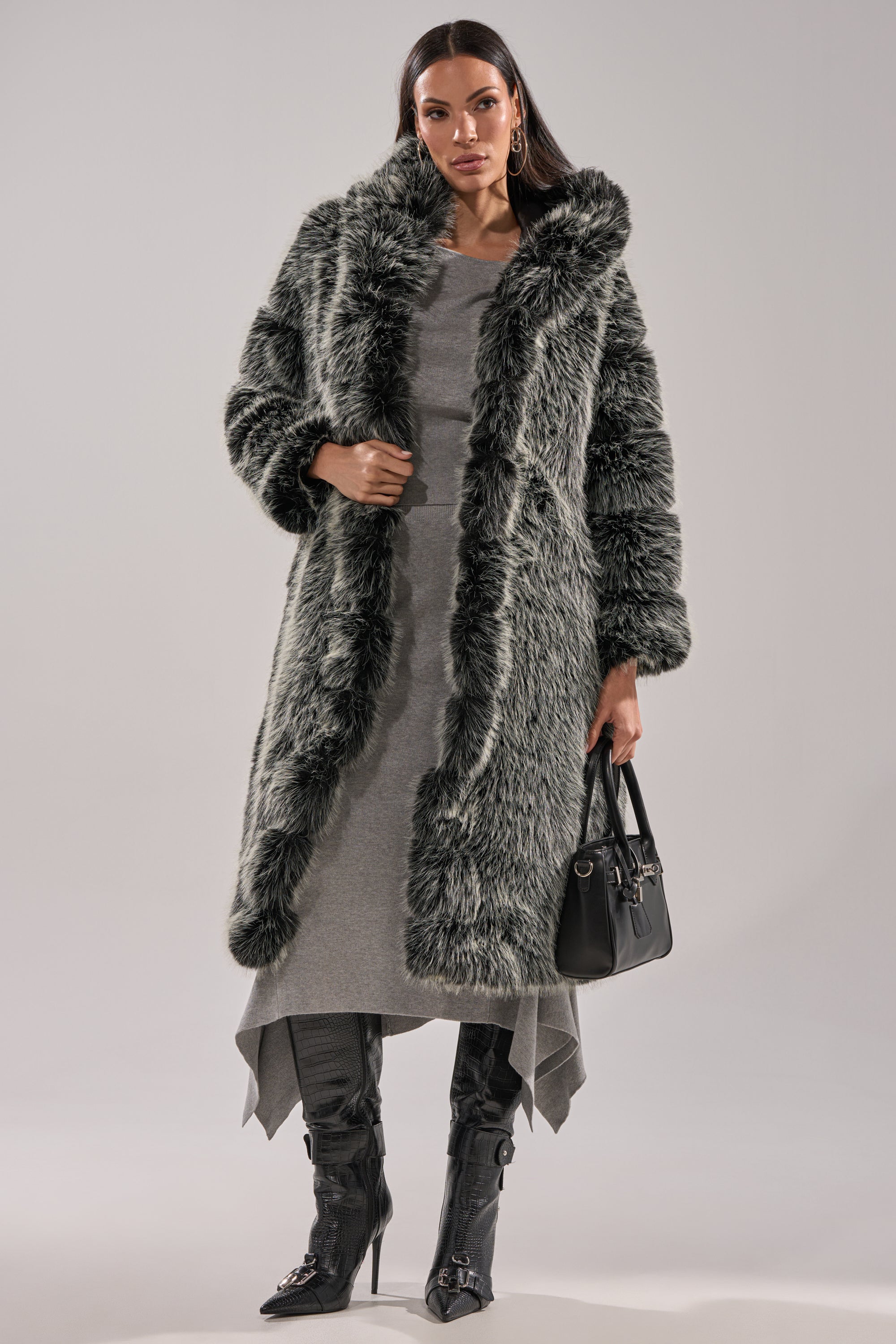 A woman in the SNOW BUNNY HOODED FAUX FUR COAT layered over a gray dress, with black high-heeled boots and a black handbag, poses confidently against a plain light background.