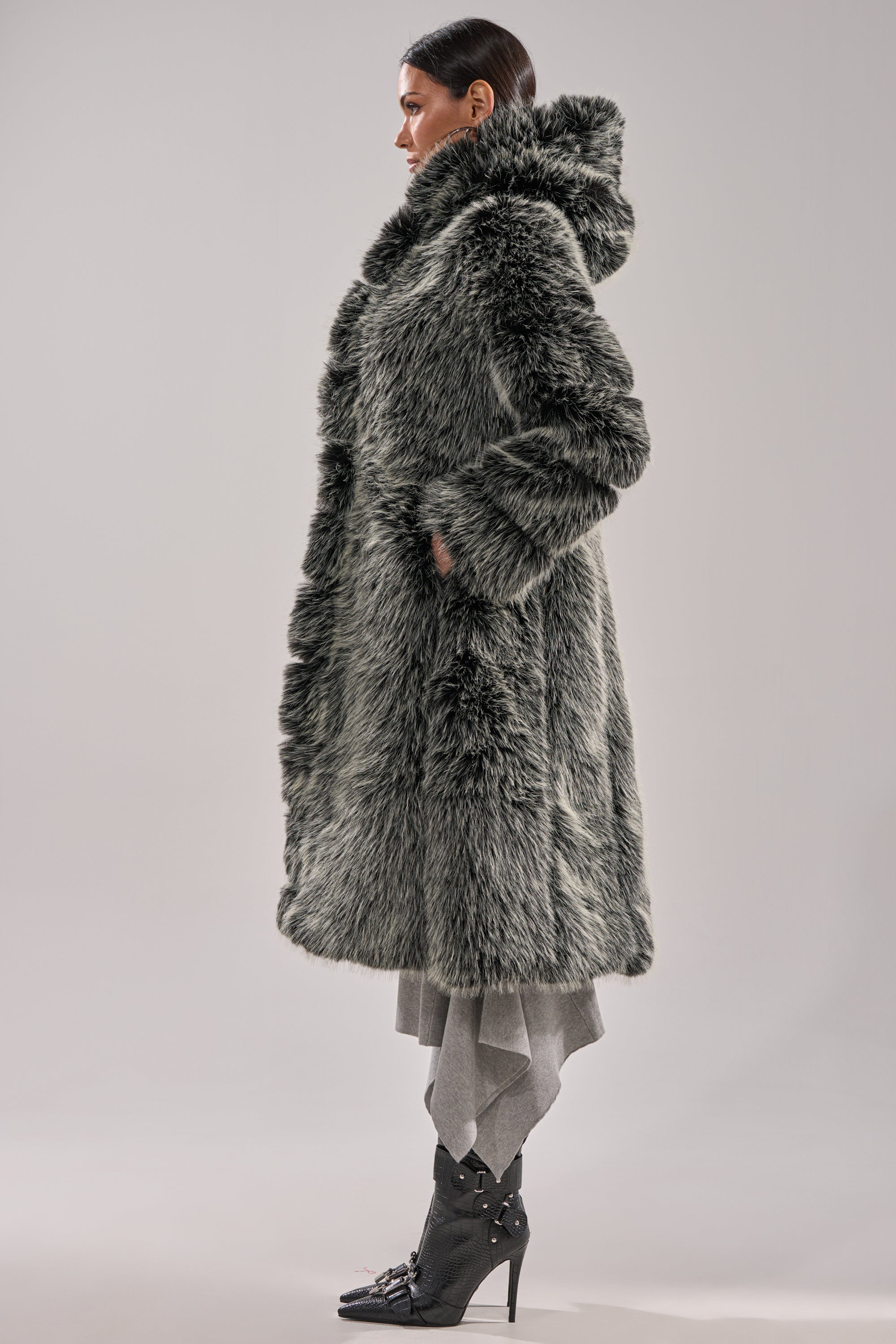 A woman in profile wears the SNOW BUNNY HOODED FAUX FUR COAT with a longline silhouette, hands in pockets. She pairs it with layered gray pants and black buckled high-heeled boots.